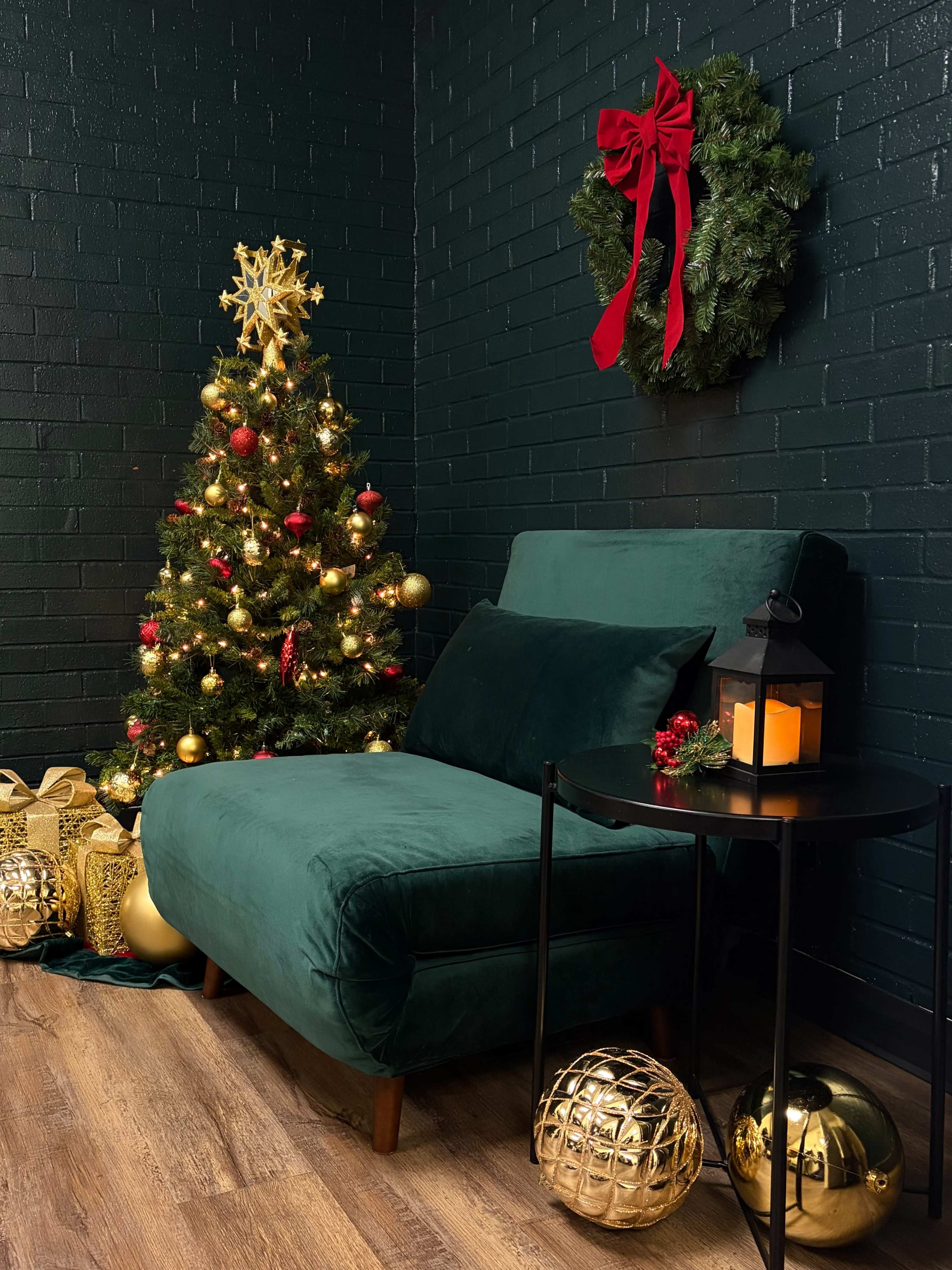 A green velvet chaise lounge is placed beside a decorated Christmas tree and a festive wreath against a dark brick wall.