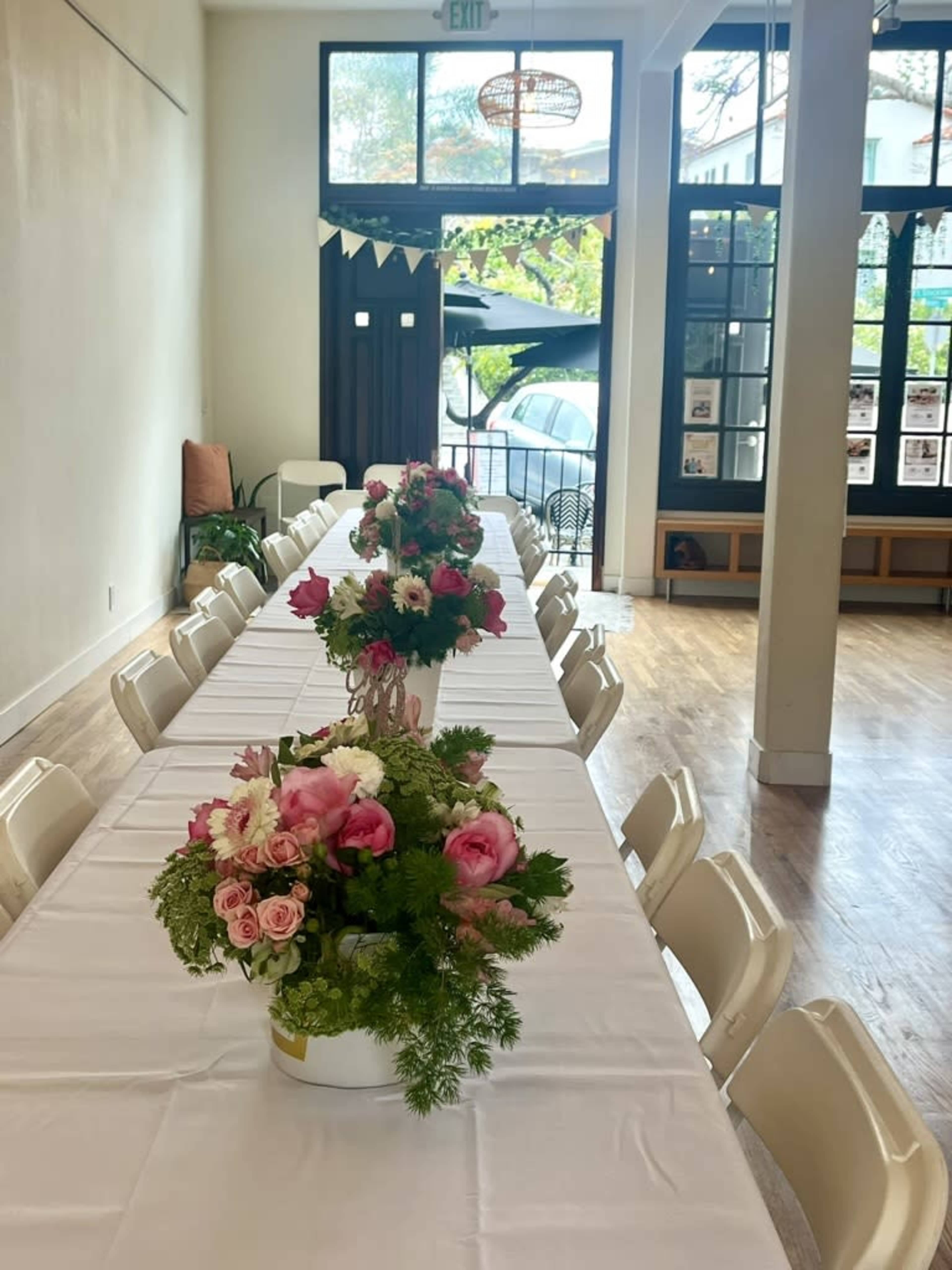 A long, white-topped dining table is set with floral centerpieces in a bright room that opens to an outdoor area.