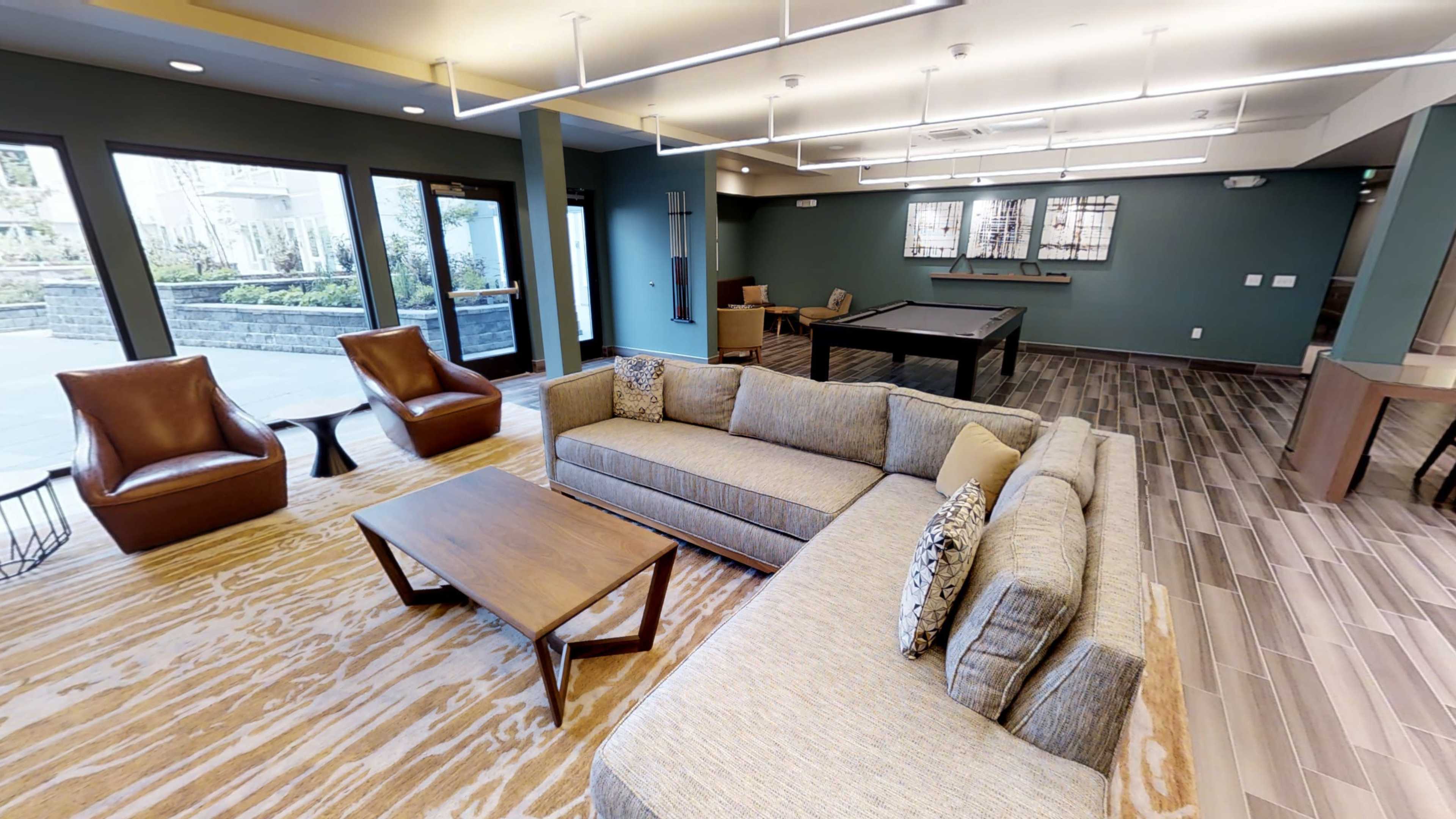 A modern lounge area features a sectional sofa, wooden coffee table, and two armchairs, with large windows overlooking a garden and a pool table in the background.