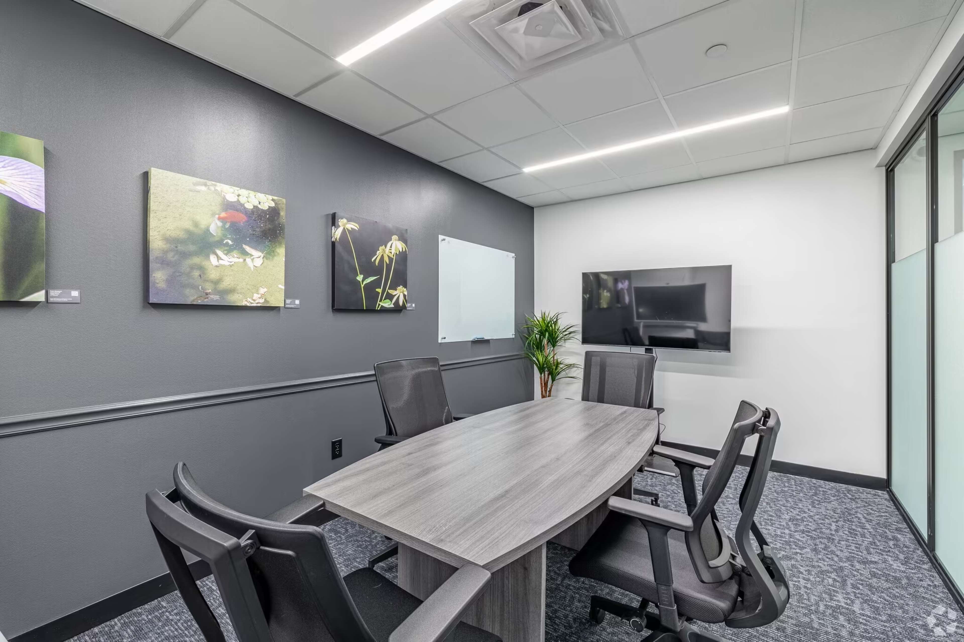 A modern meeting room features a large rectangular table surrounded by black chairs, framed artwork on the walls, a flat-screen TV, and a whiteboard.