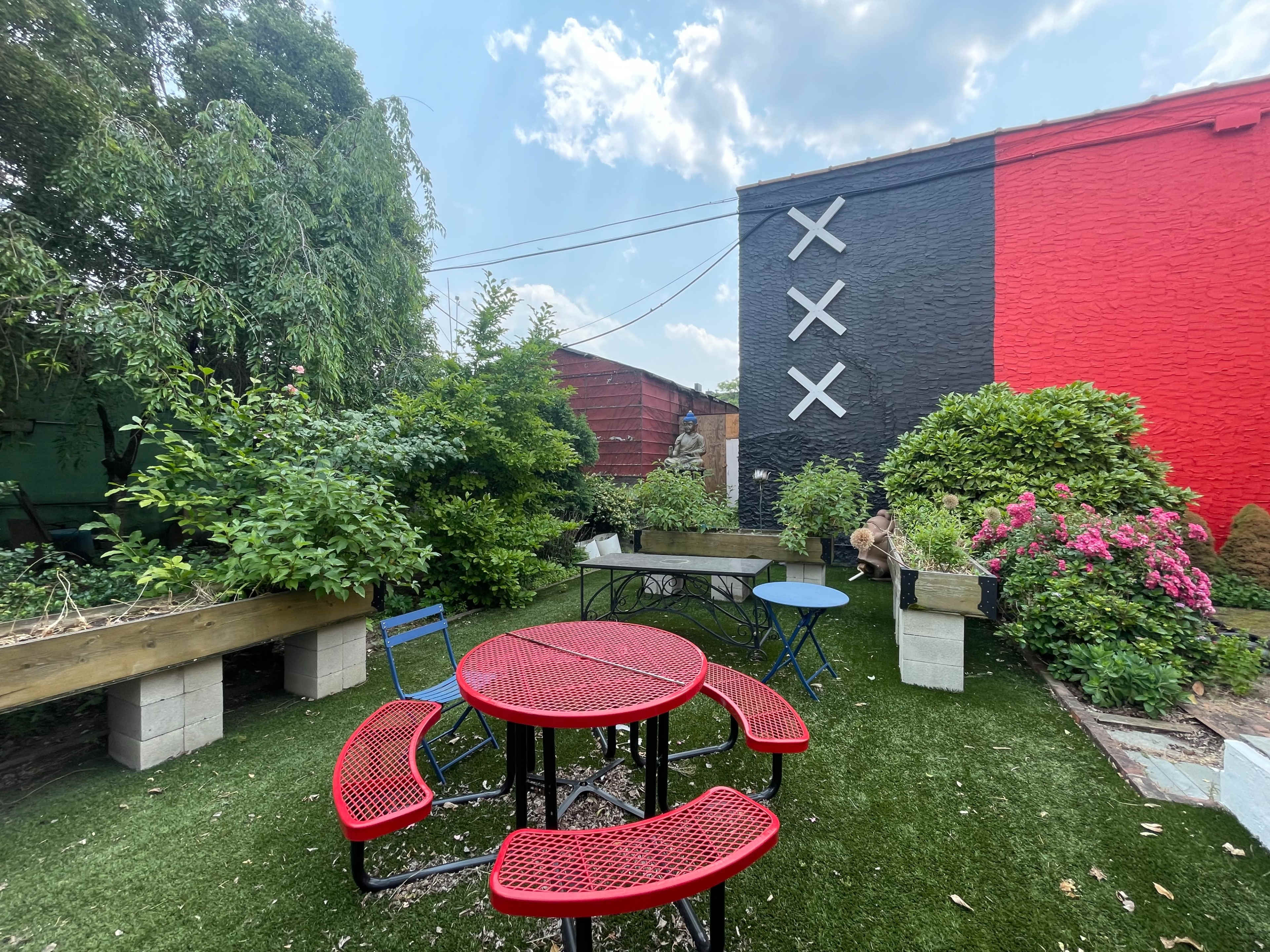 A landscaped outdoor area with red picnic tables, green plants, and a mural featuring black and red walls adorned with white cross symbols.