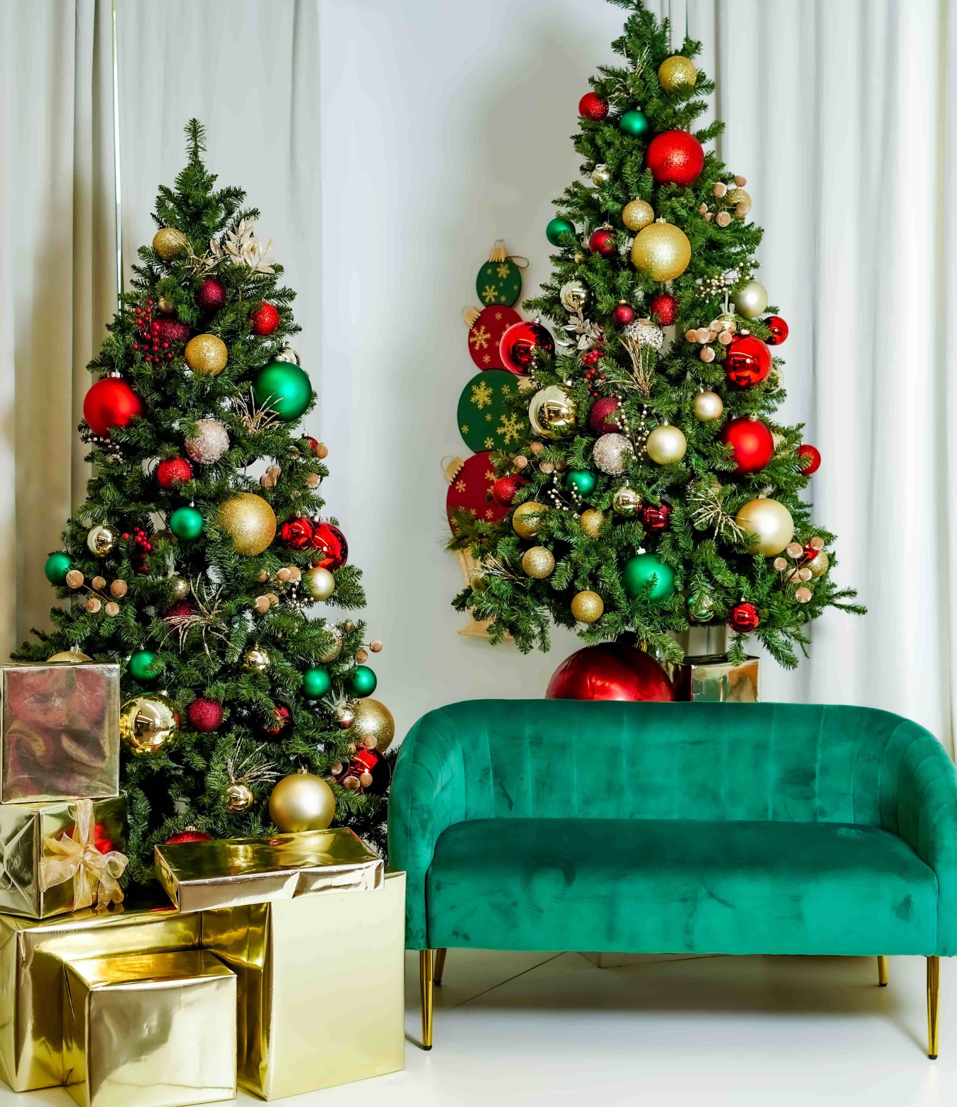 Two Christmas trees adorned with ornaments stand on either side of a green velvet sofa, while gold and silver wrapped gifts are arranged nearby.