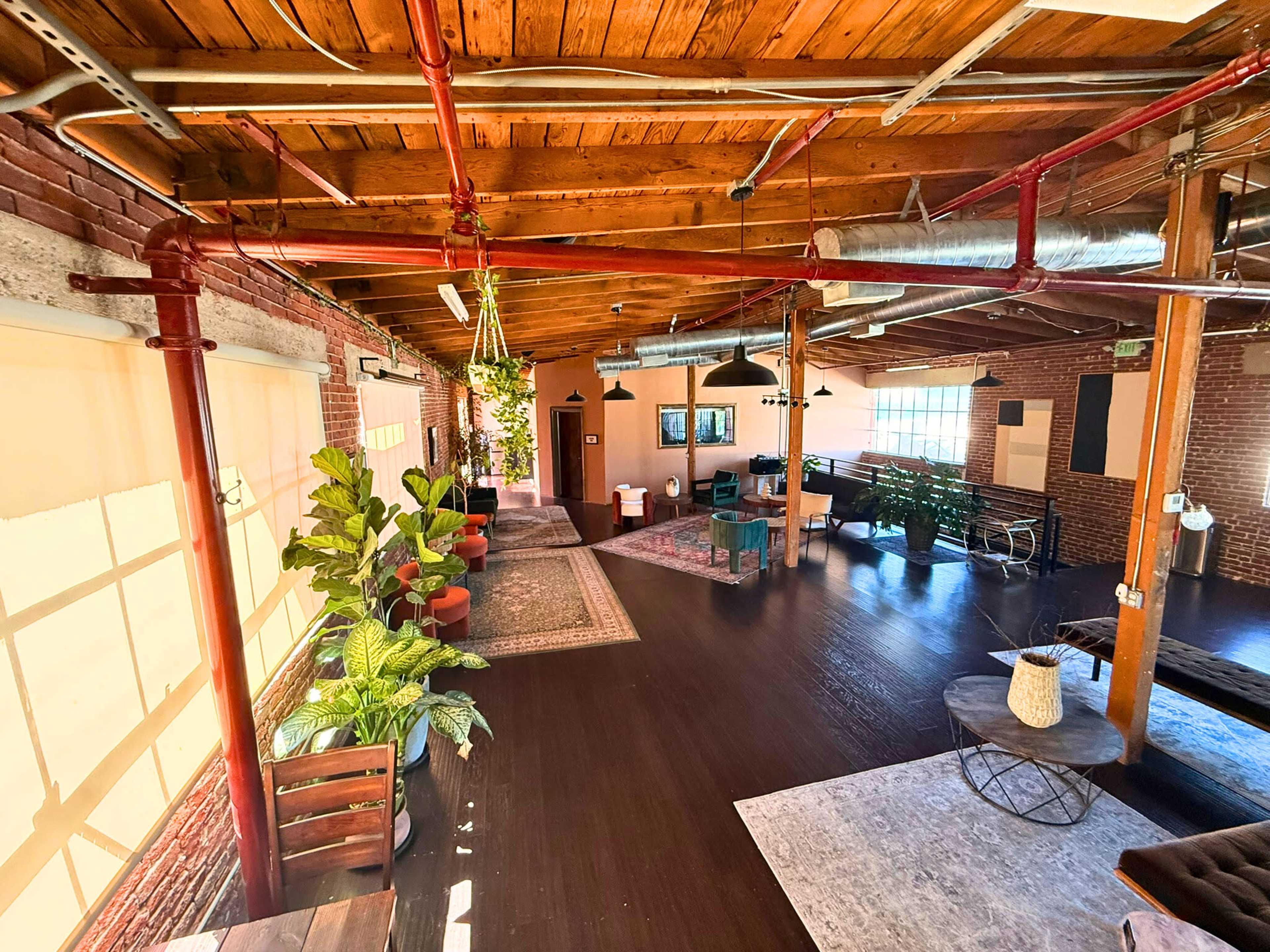 The image shows a spacious interior with wooden beams, potted plants, and various seating areas, illuminated by natural light coming through large windows.