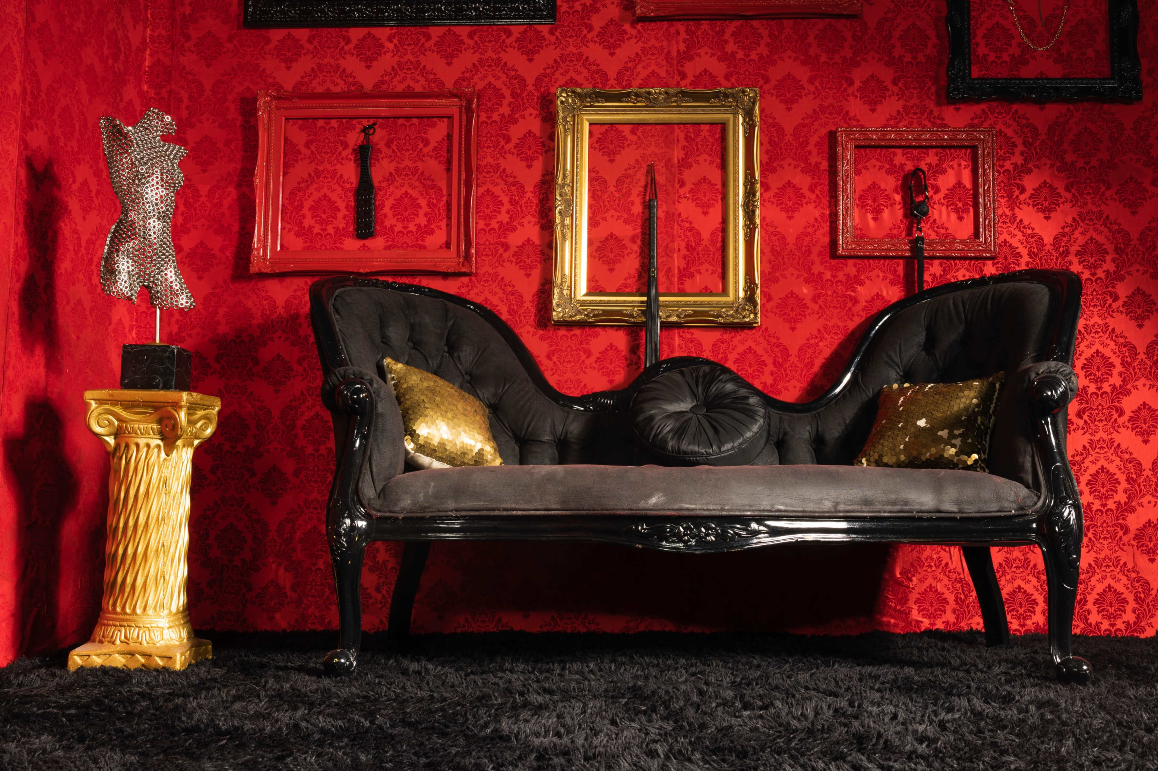 A black vintage loveseat against a red wallpaper background, surrounded by ornate gold and black picture frames and a decorative pedestal holding a shiny, sculptural figure.