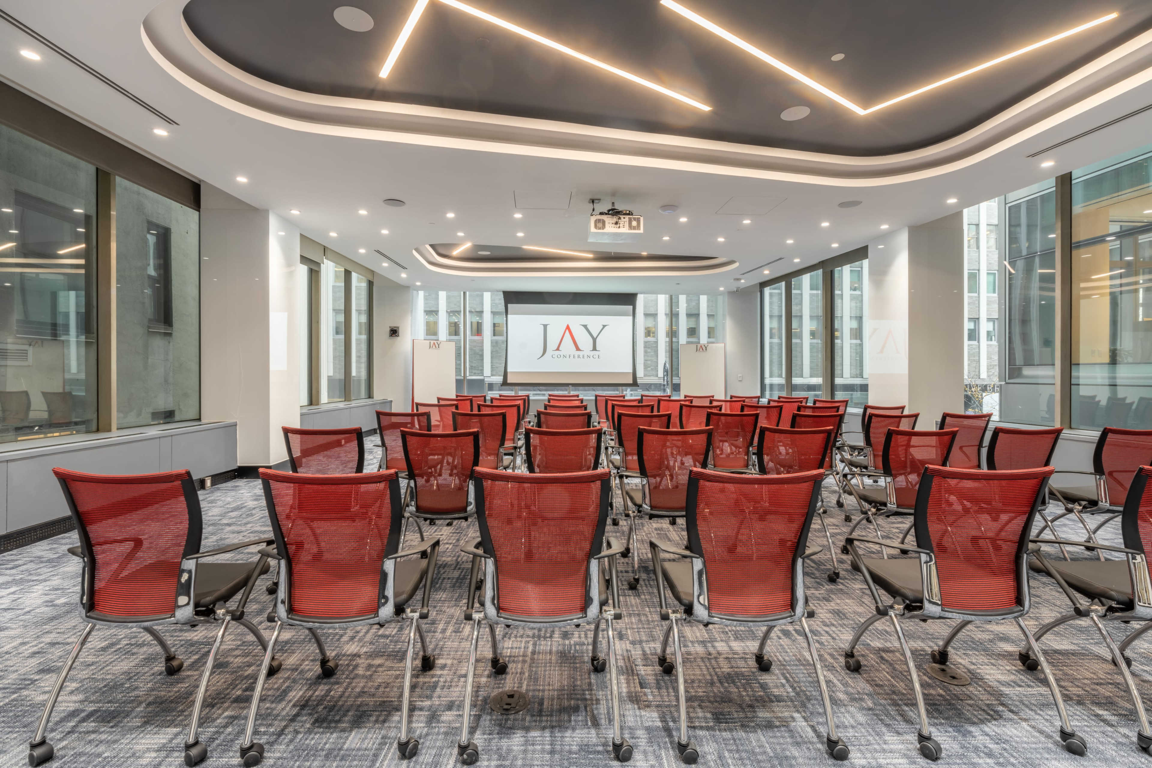 A conference room with rows of red chairs facing a large screen displaying the logo "JAY".