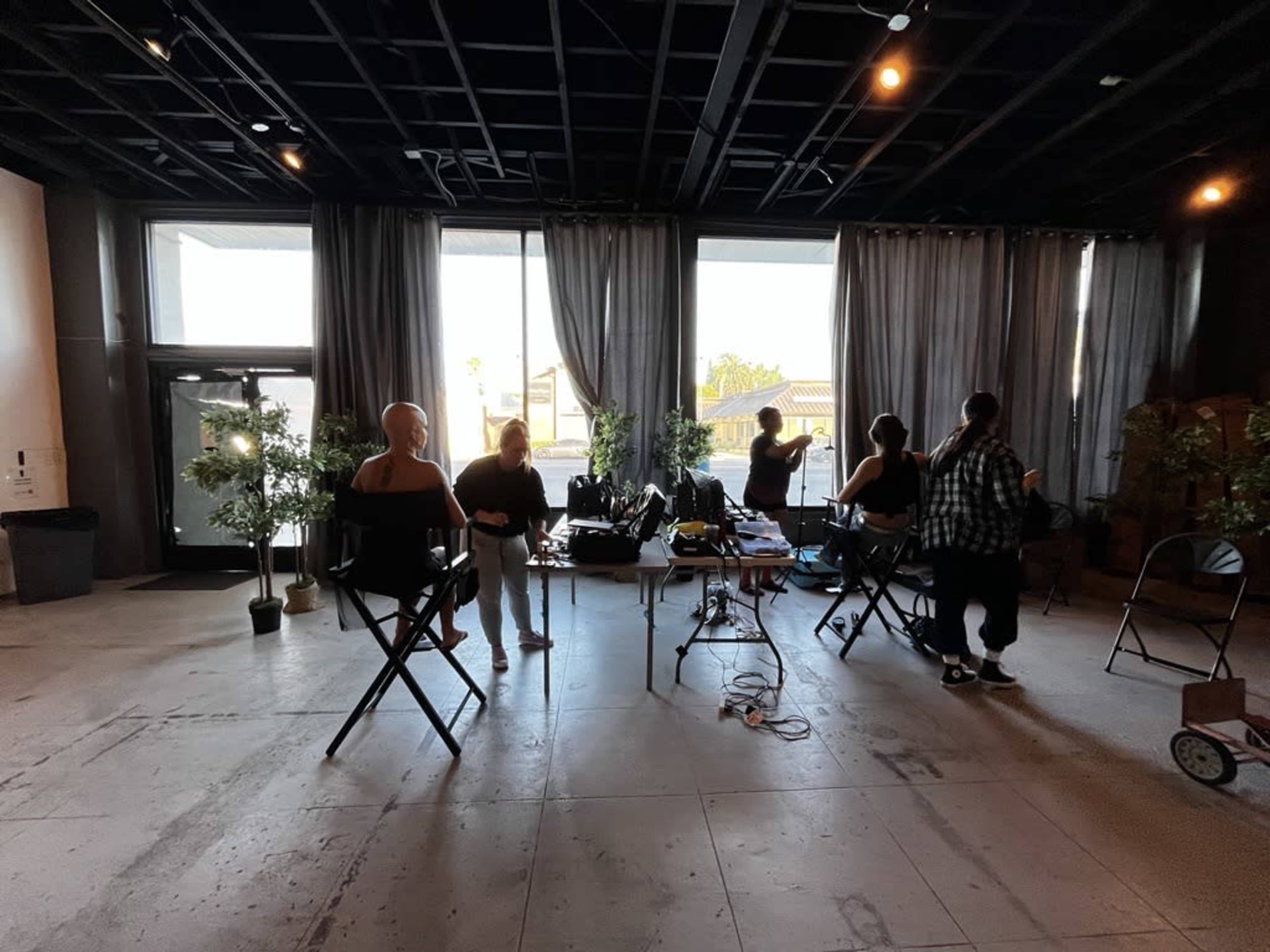 A group of individuals is working in a spacious studio with tall windows and various equipment set up for a production or photoshoot.