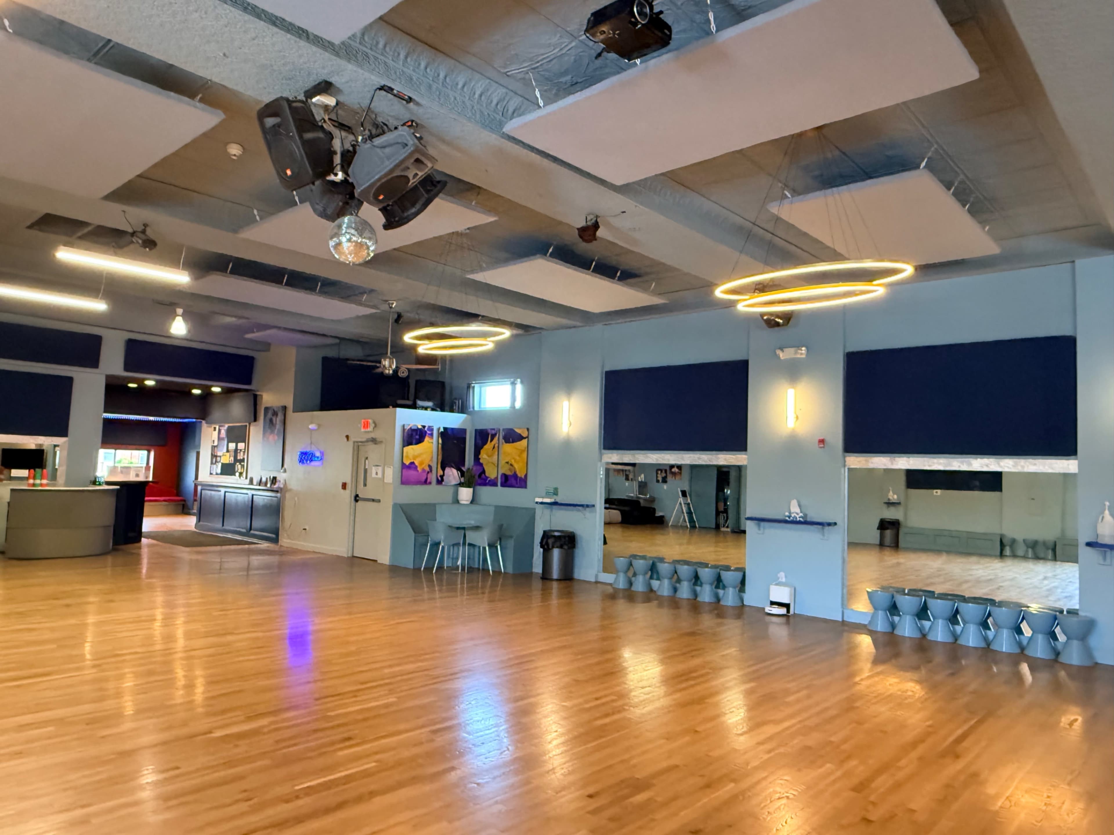 The image shows a large, empty dance studio with wooden flooring, large mirrors on one wall, and modern lighting fixtures.