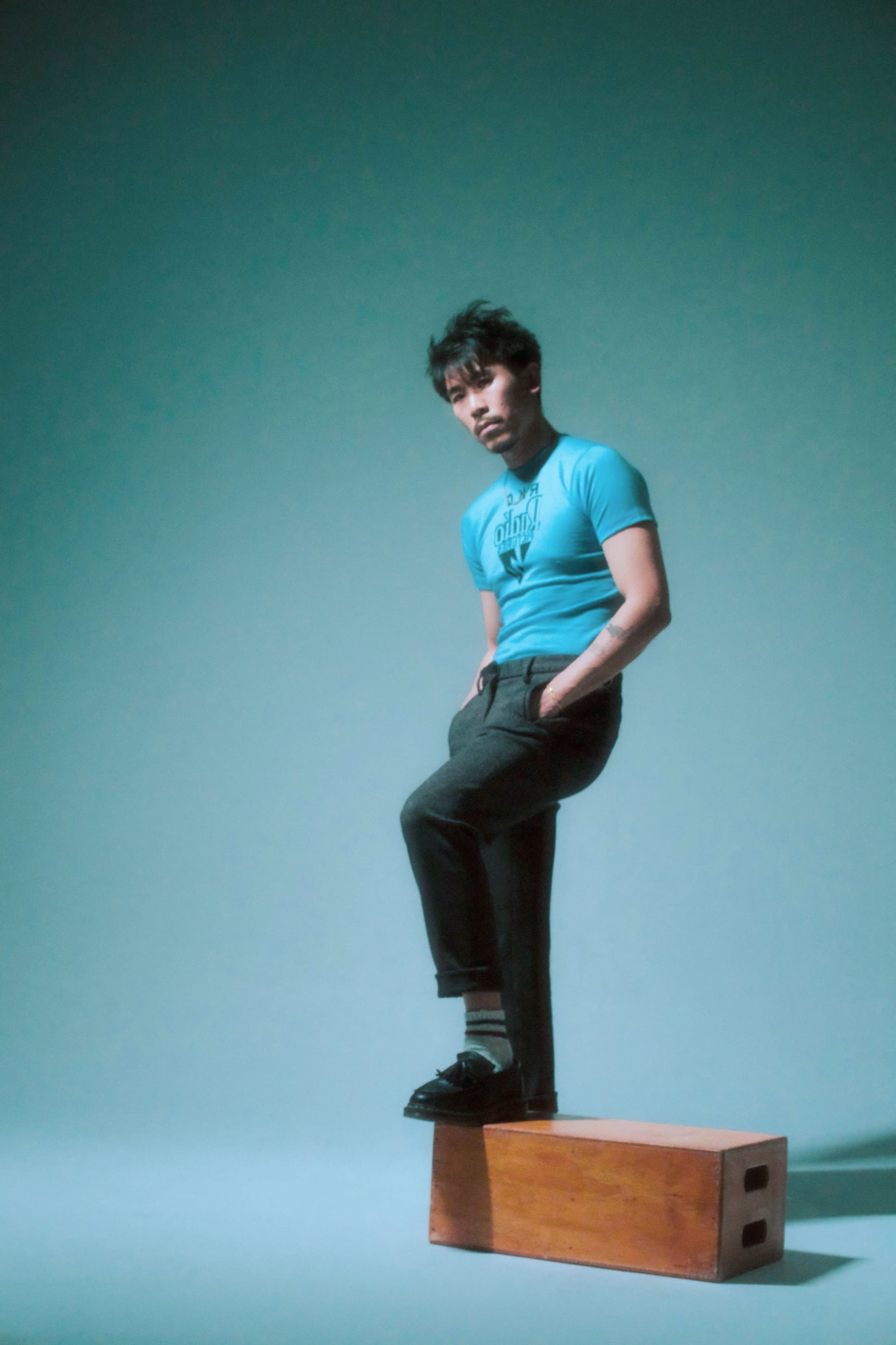 A man stands on a wooden block, wearing a blue shirt and gray trousers, against a soft blue backdrop.