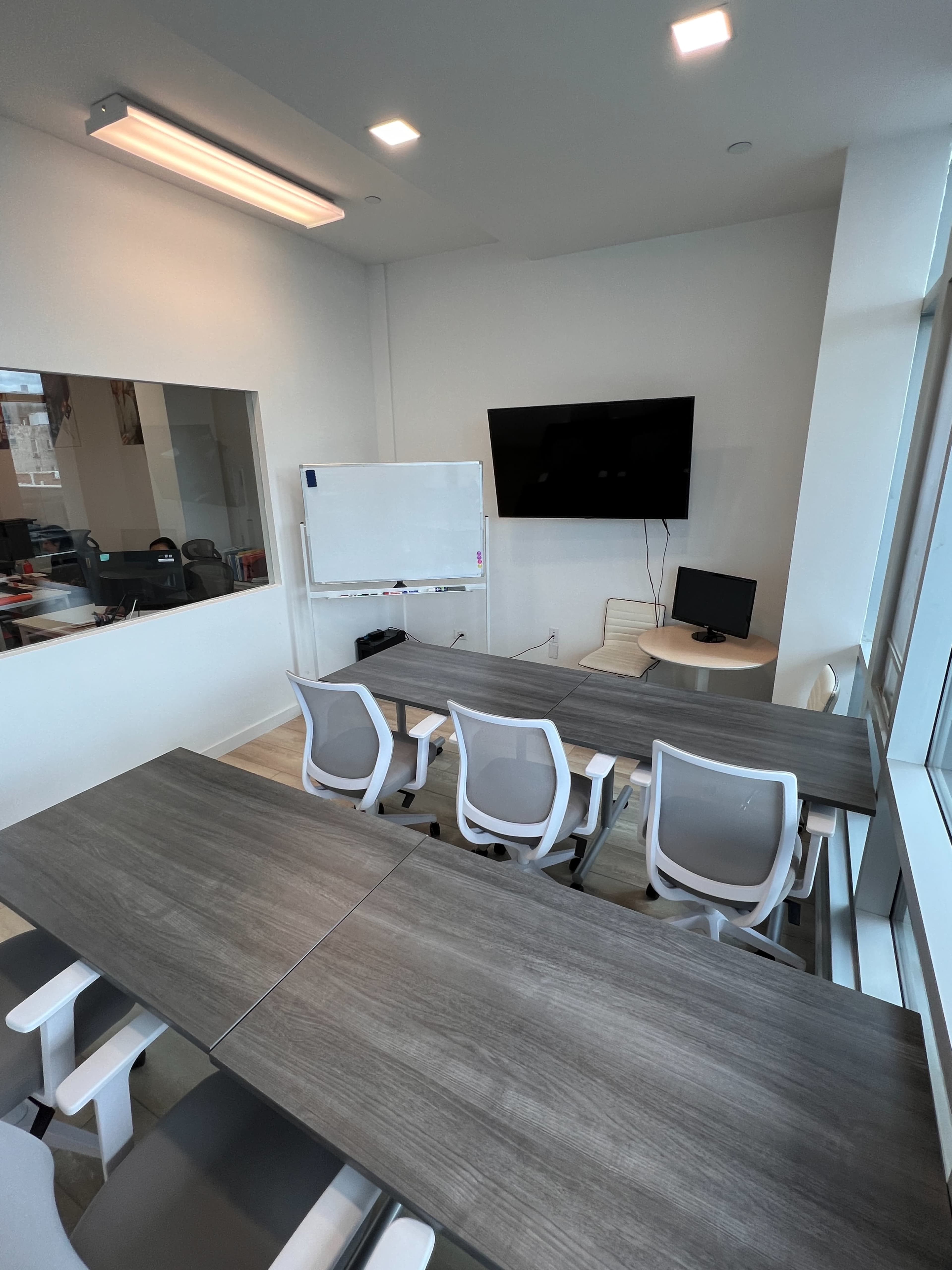 A modern conference room features a large table with several chairs, a whiteboard, a television on the wall, and a computer workstation in the corner.