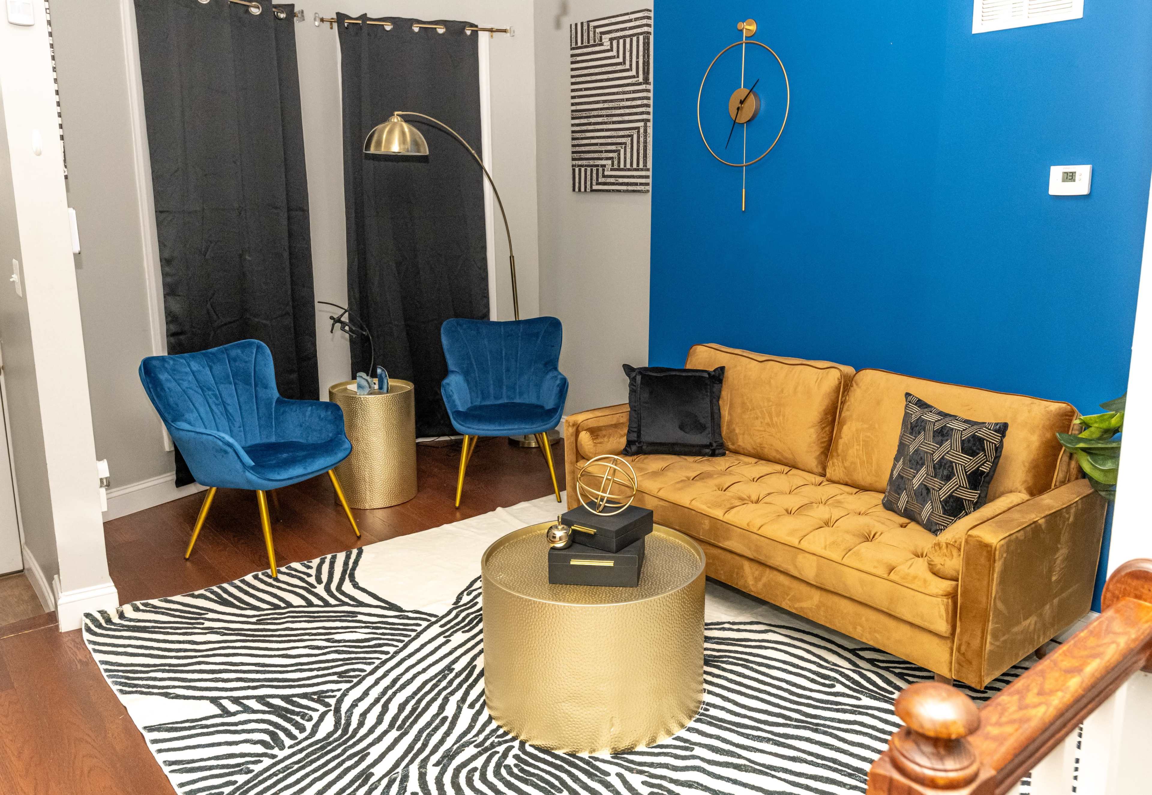 The scene features a cozy living room with a gold tufted sofa, two blue accent chairs, a round gold coffee table, and a zebra-patterned rug against a blue wall.
