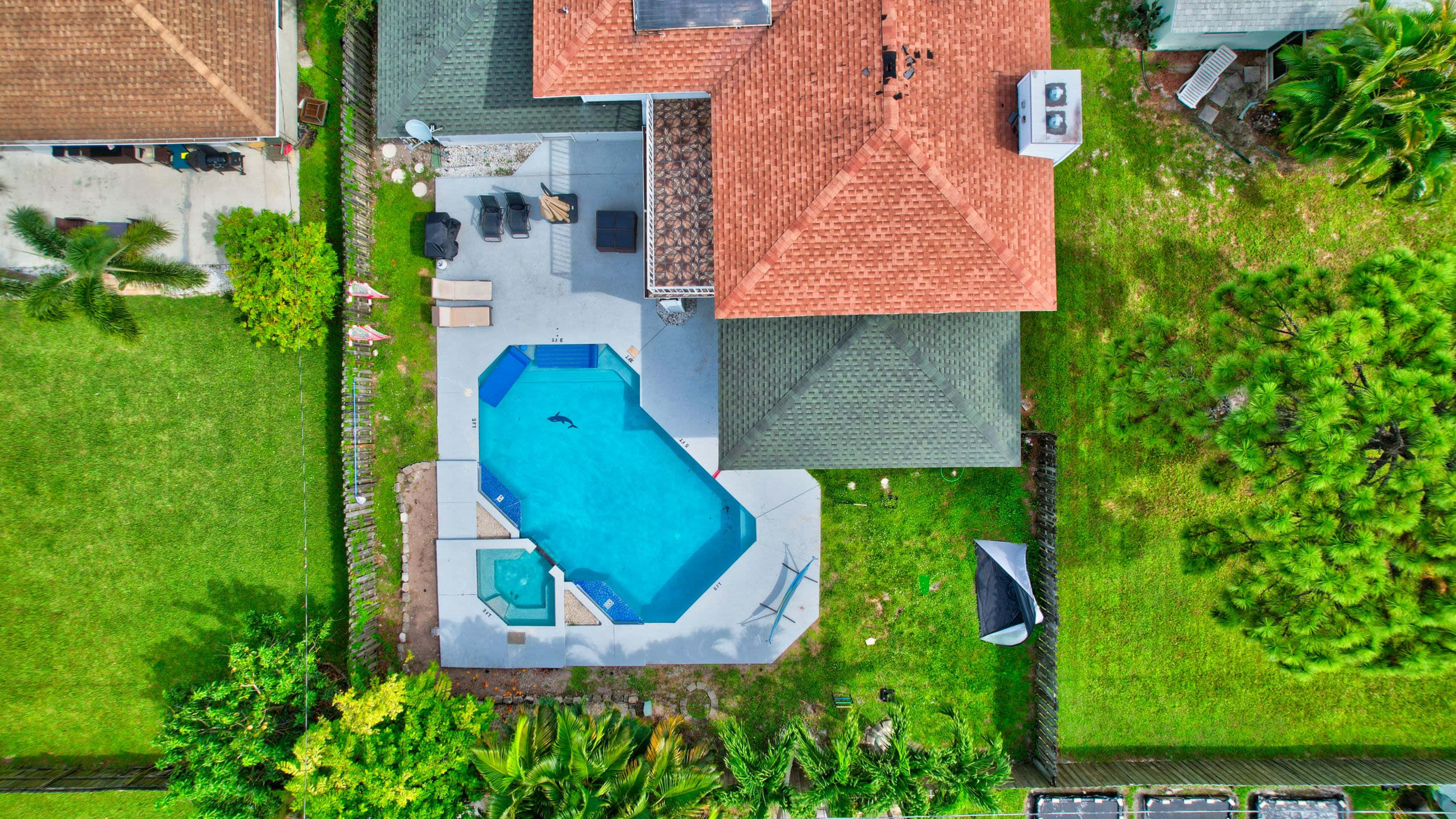 An aerial view shows a backyard with a swimming pool, surrounding patio, and two nearby houses set in green landscaping.