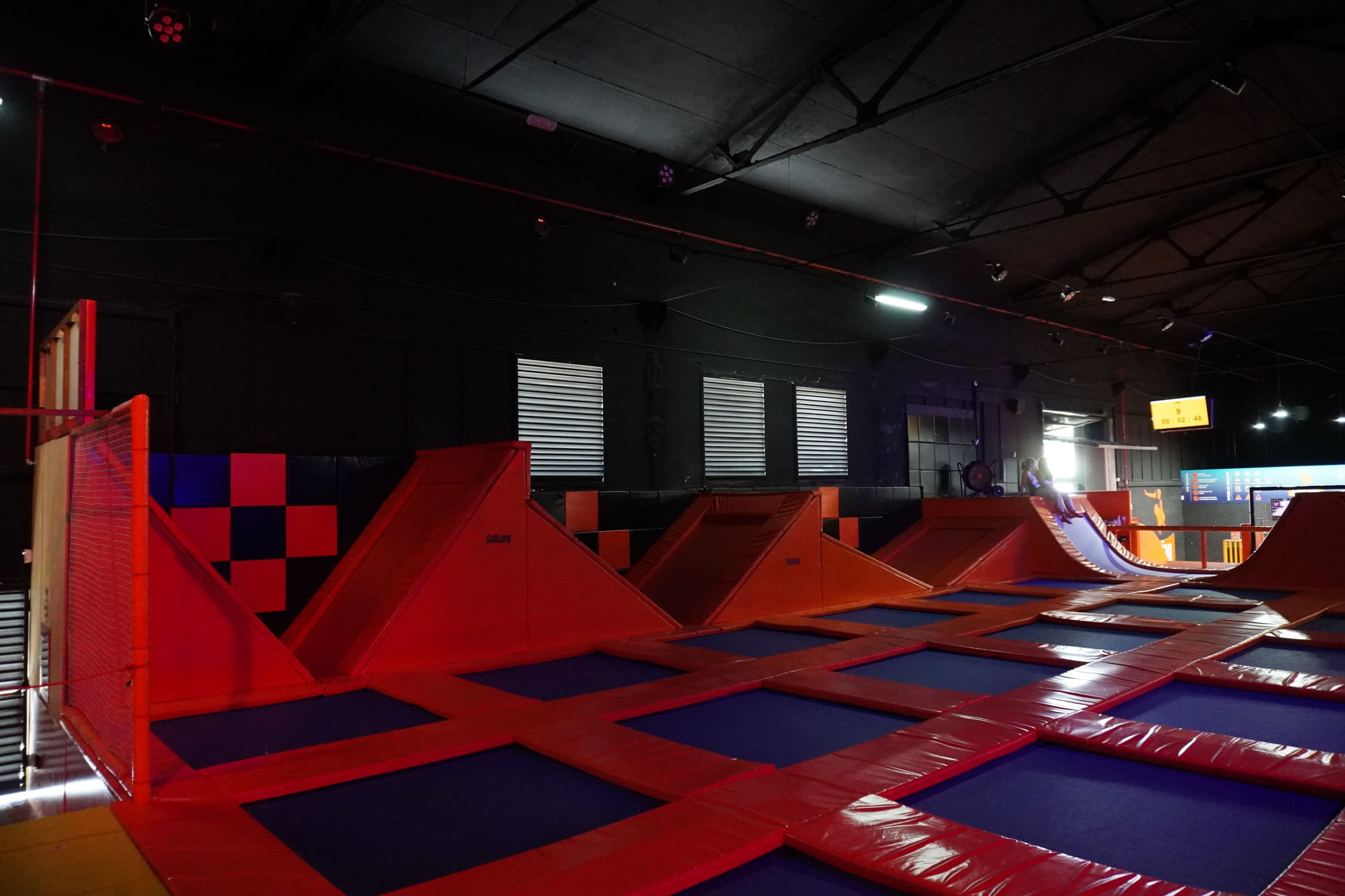A trampoline park with several orange and blue trampolines and angled ramps in a dark indoor setting.