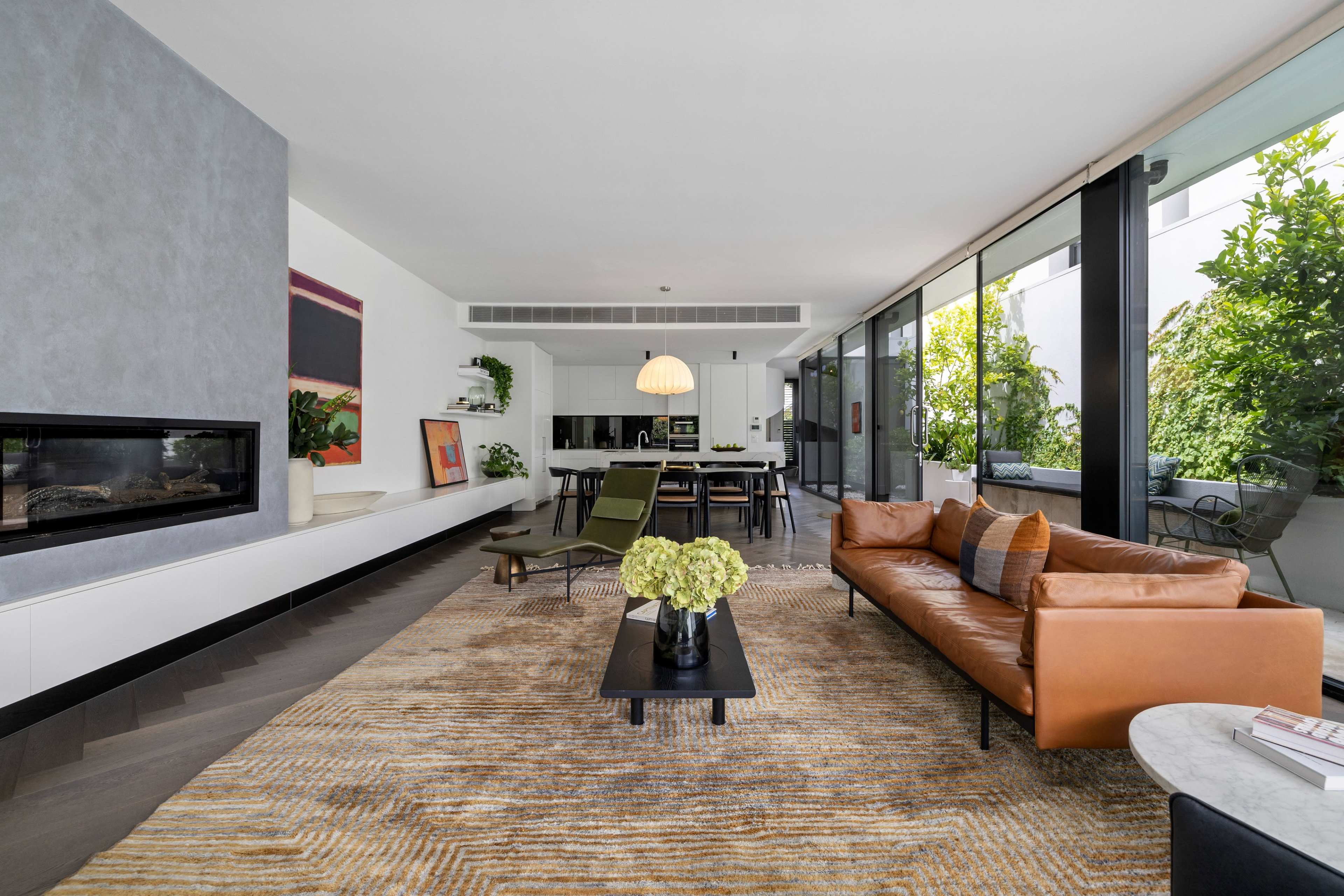 A modern living room featuring a brown leather sofa, a coffee table with flowers, and large windows overlooking greenery.