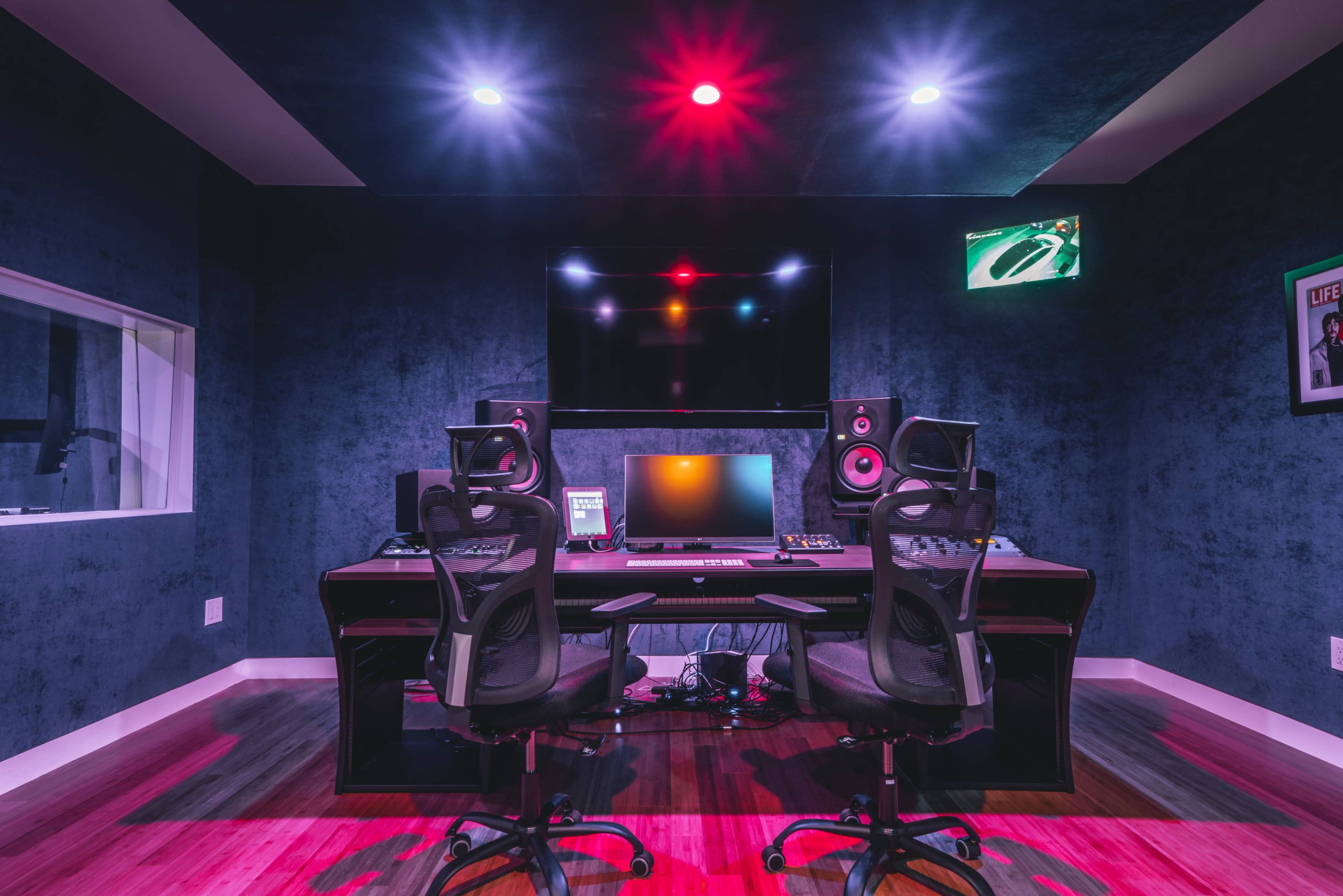 A modern music studio featuring a central desk equipped with audio equipment, two black chairs, and colorful LED lights on the ceiling.