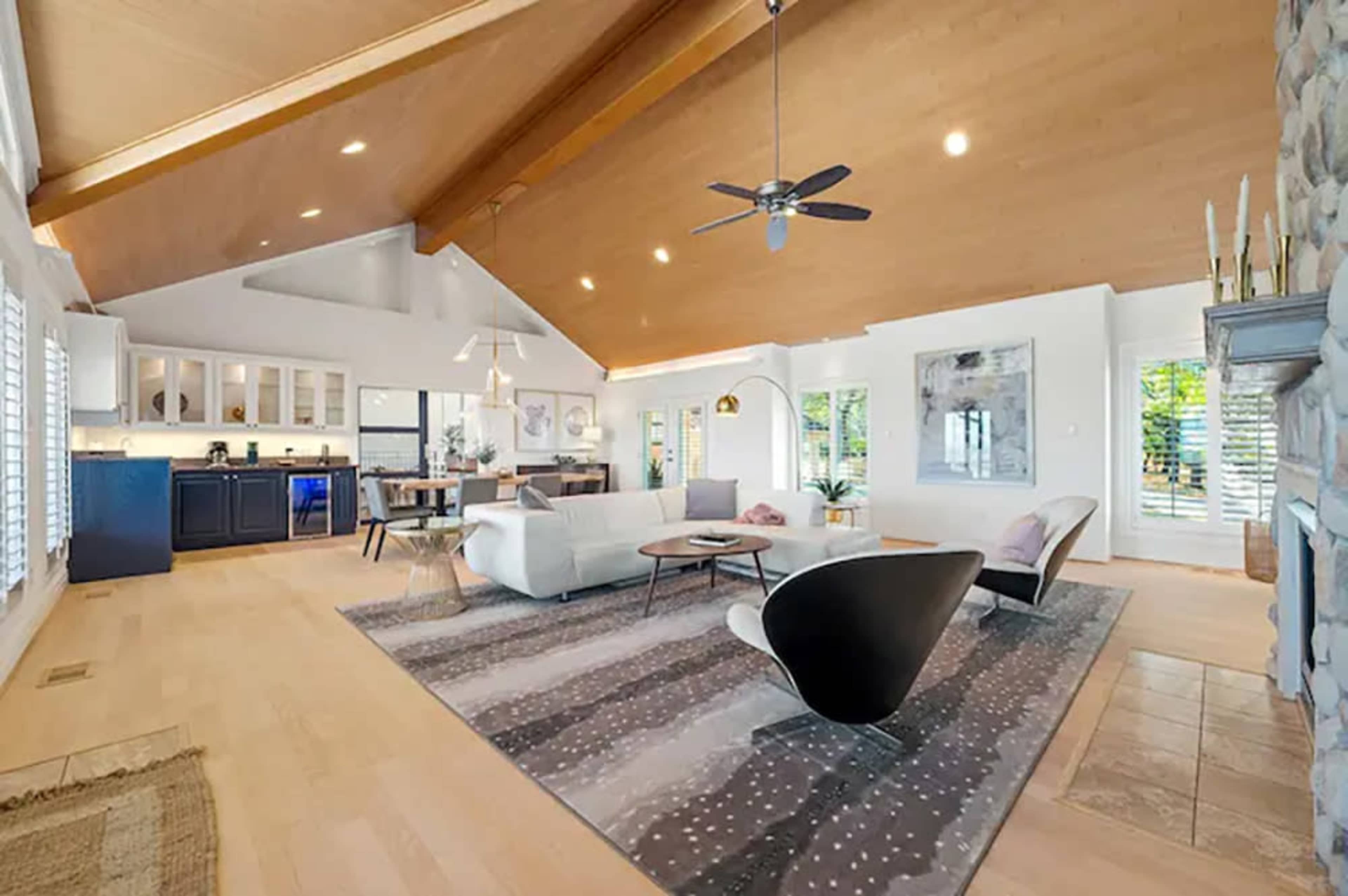 The image shows a spacious living room with a vaulted wooden ceiling, modern furniture, and large windows that allow natural light to enter the space.