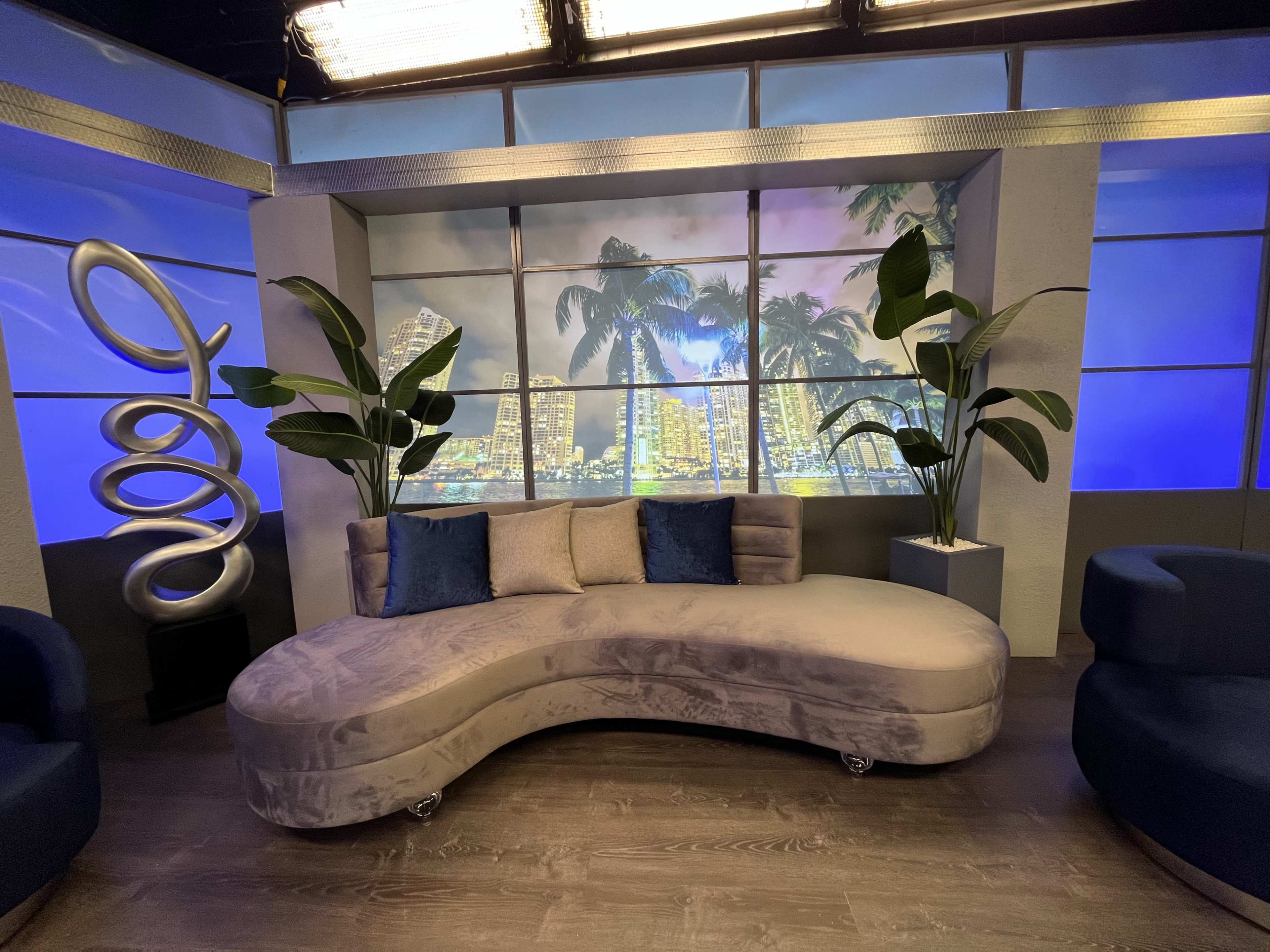A curved gray sofa is positioned in front of a backdrop of city skyscrapers and palm trees, illuminated by blue lighting.