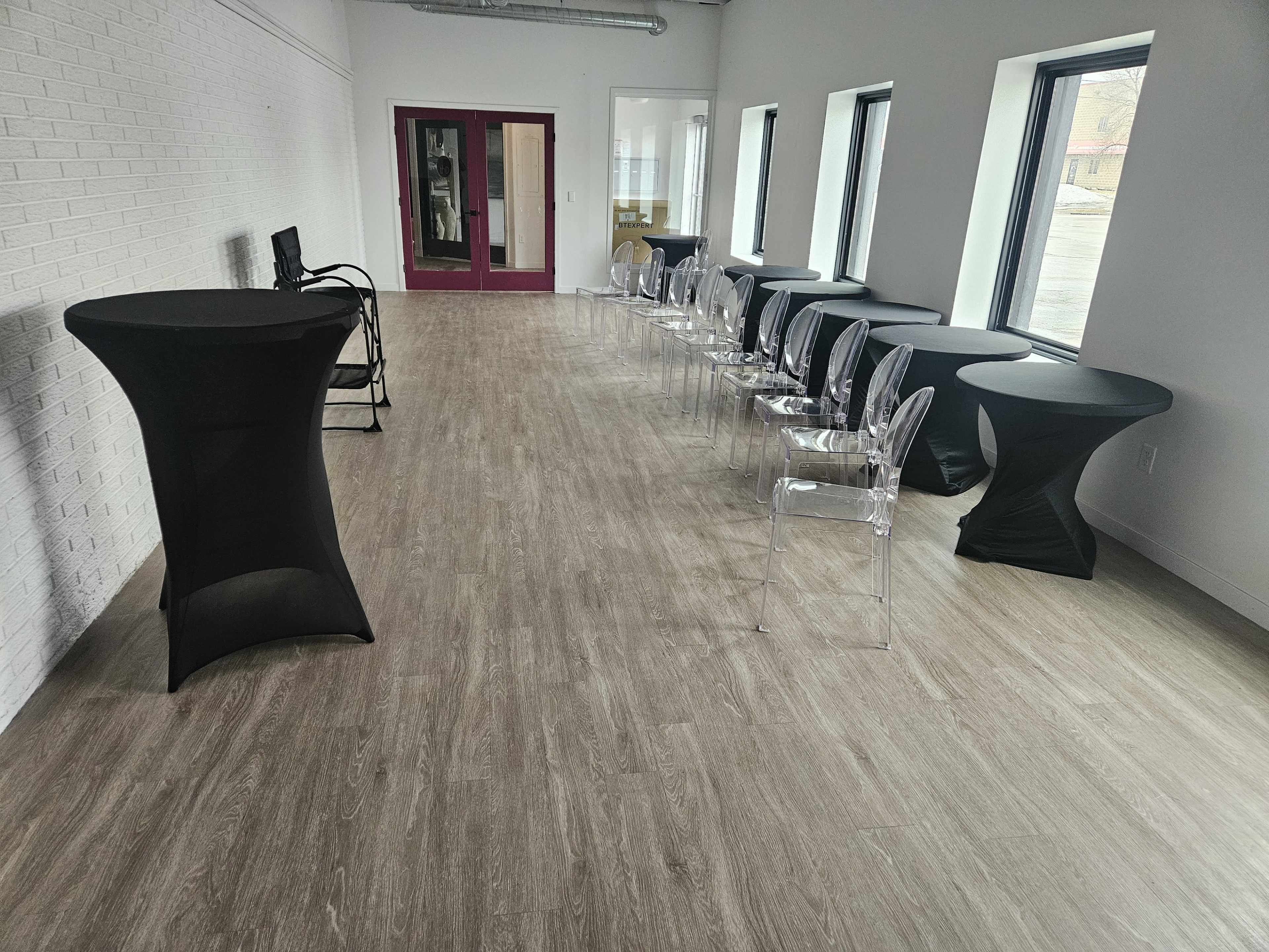The image shows a spacious room with light-colored flooring, featuring several black and transparent chairs aligned along one side and two round tables on the opposite side.