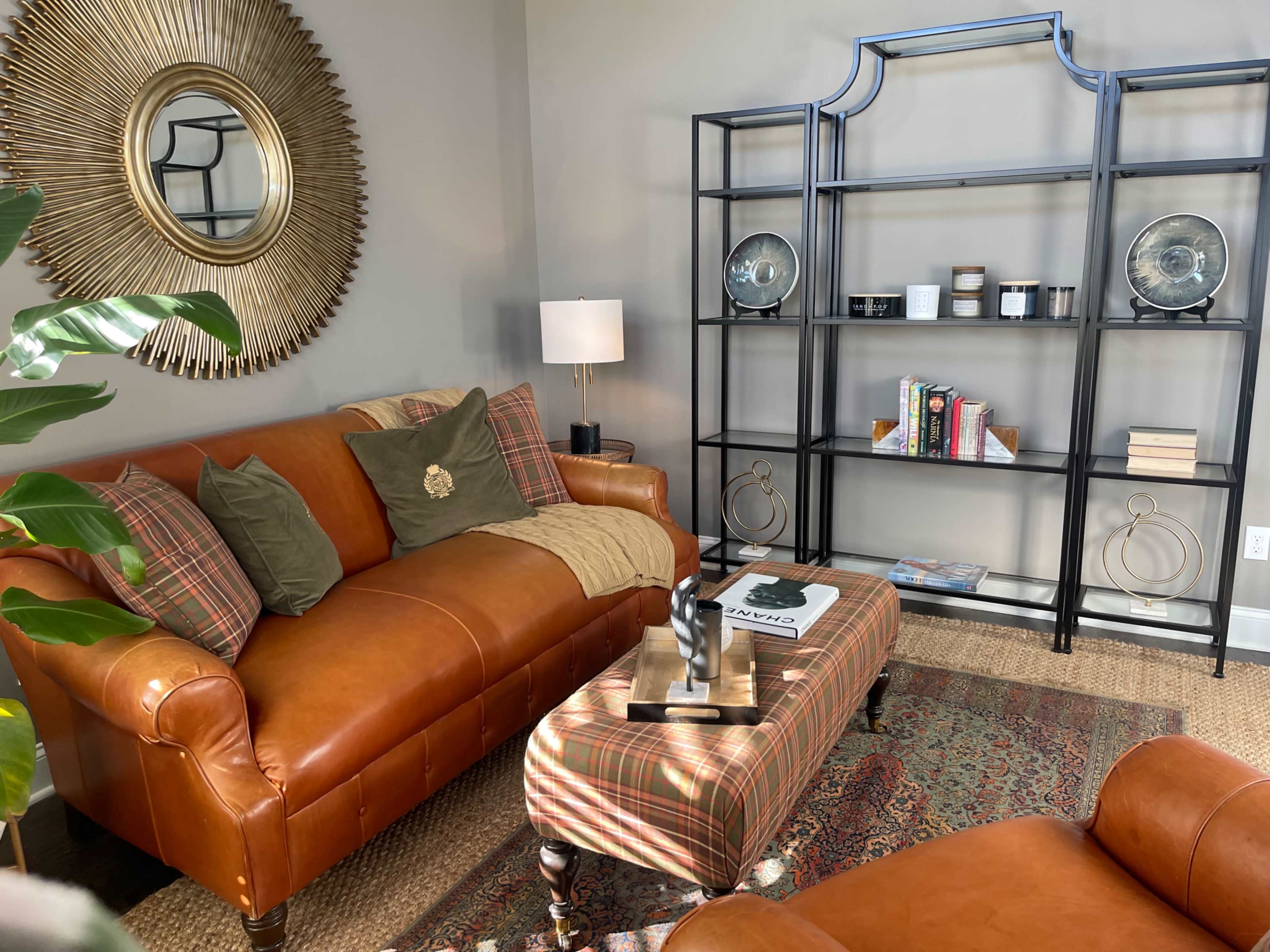 A cozy living room with a brown leather sofa, a patterned ottoman, a bookshelf, and a large circular mirror.