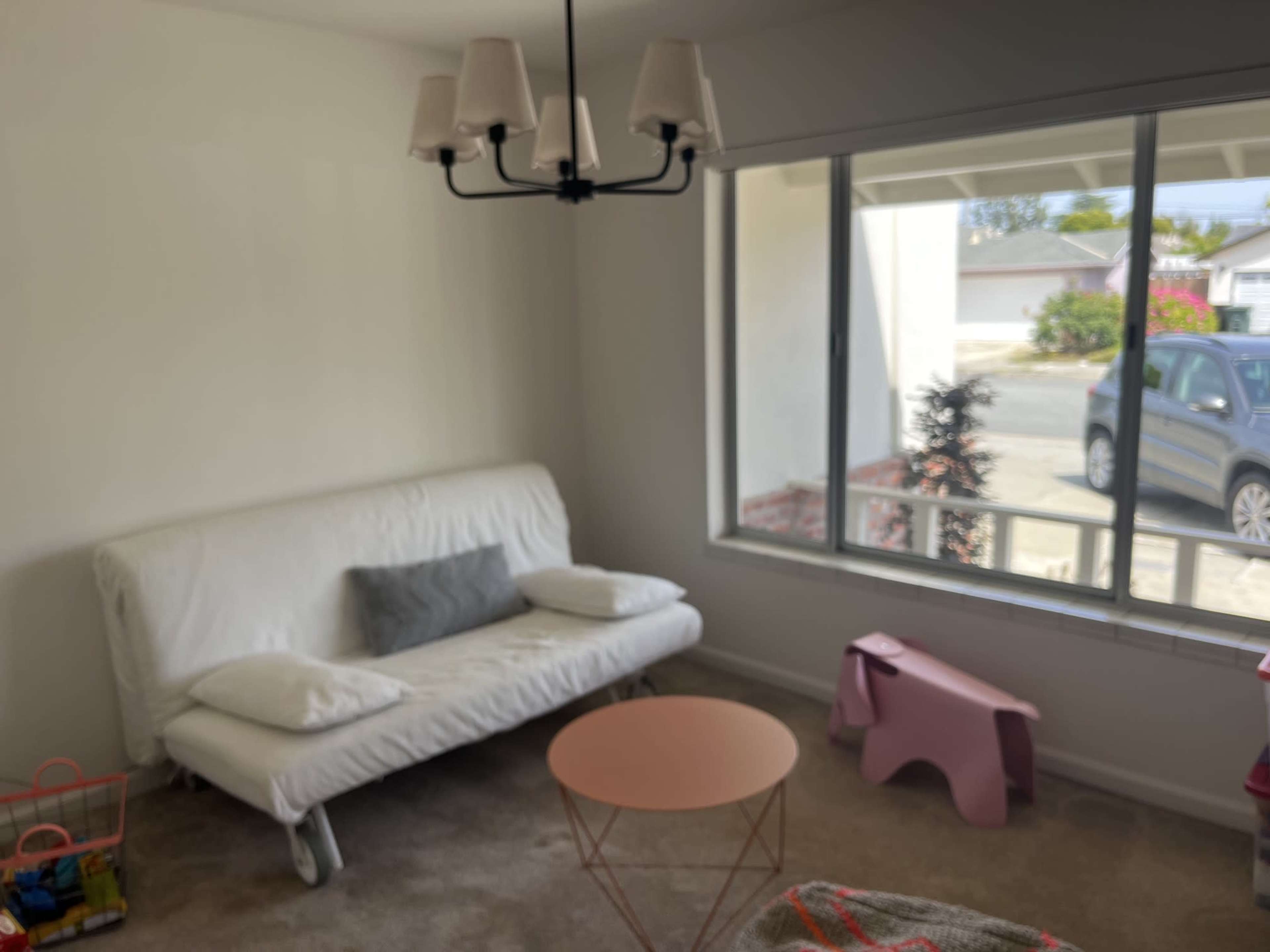 The image shows a bright room with a white futon, a pink coffee table, and a large window overlooking a driveway and street.