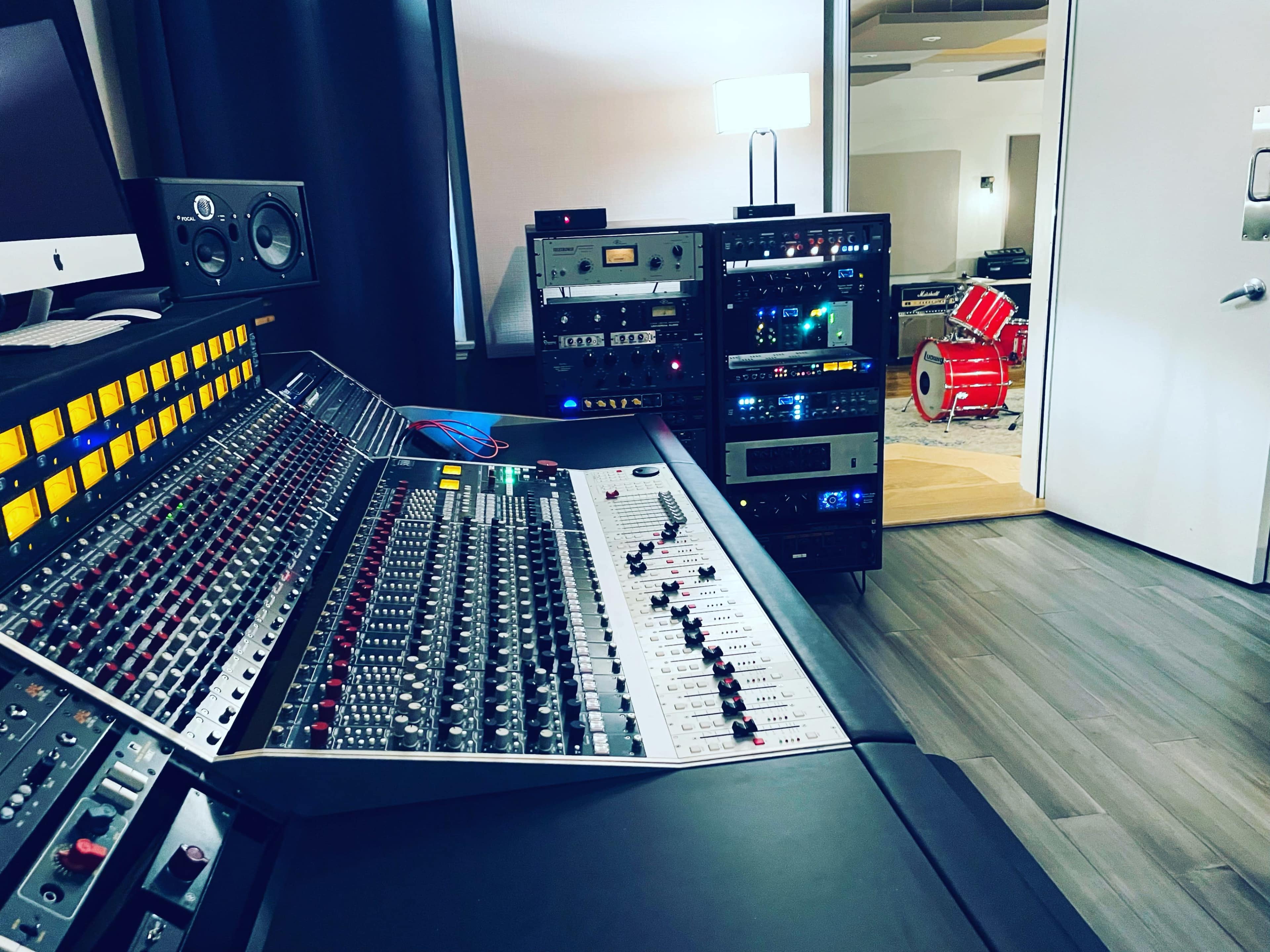 A music studio featuring a large mixing console, various audio equipment racks, and a glimpse of a drum set in an adjacent room.