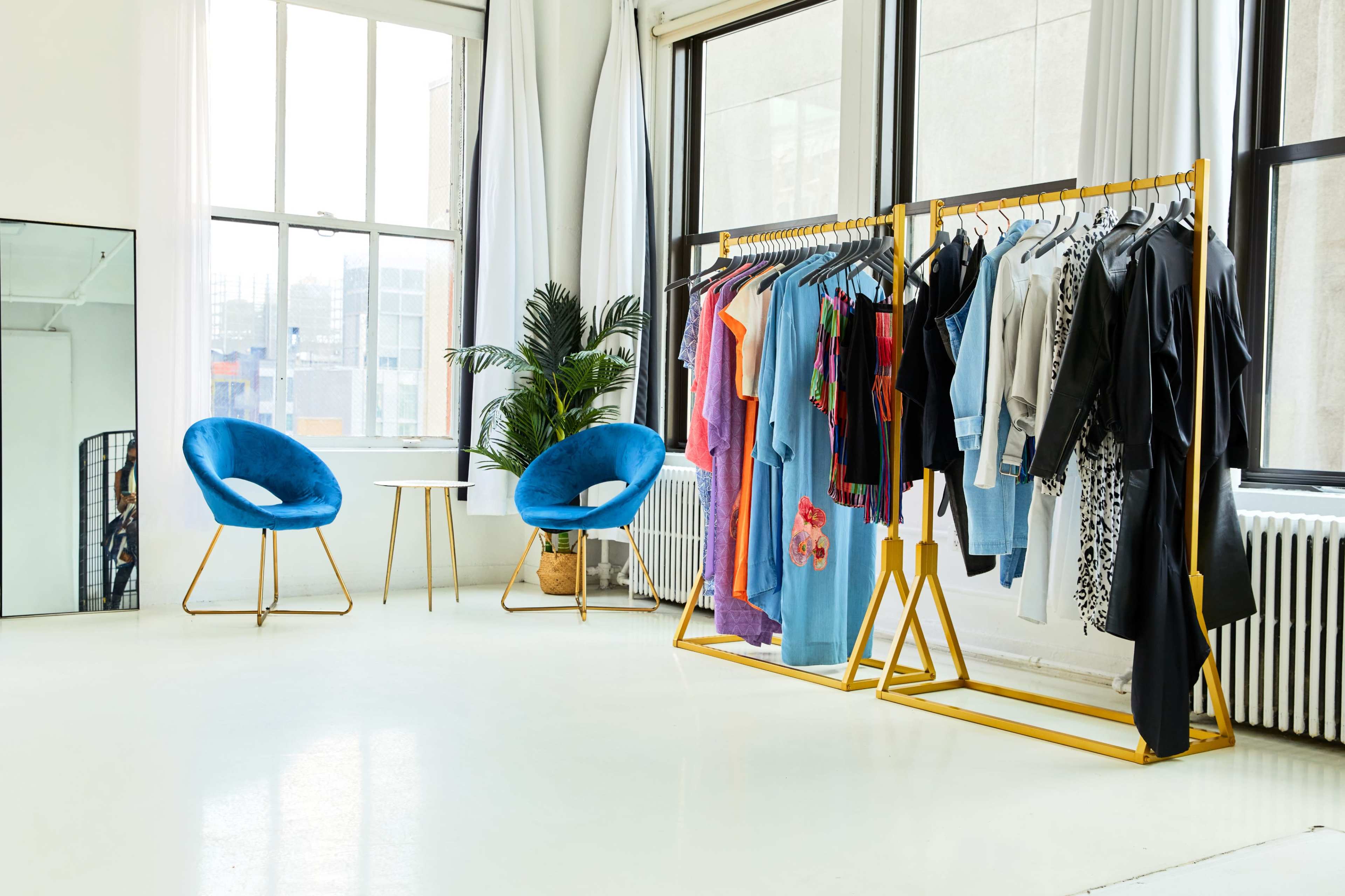 The image shows a modern clothing showroom with a clothing rack displaying various garments, two blue chairs, and large windows providing natural light.
