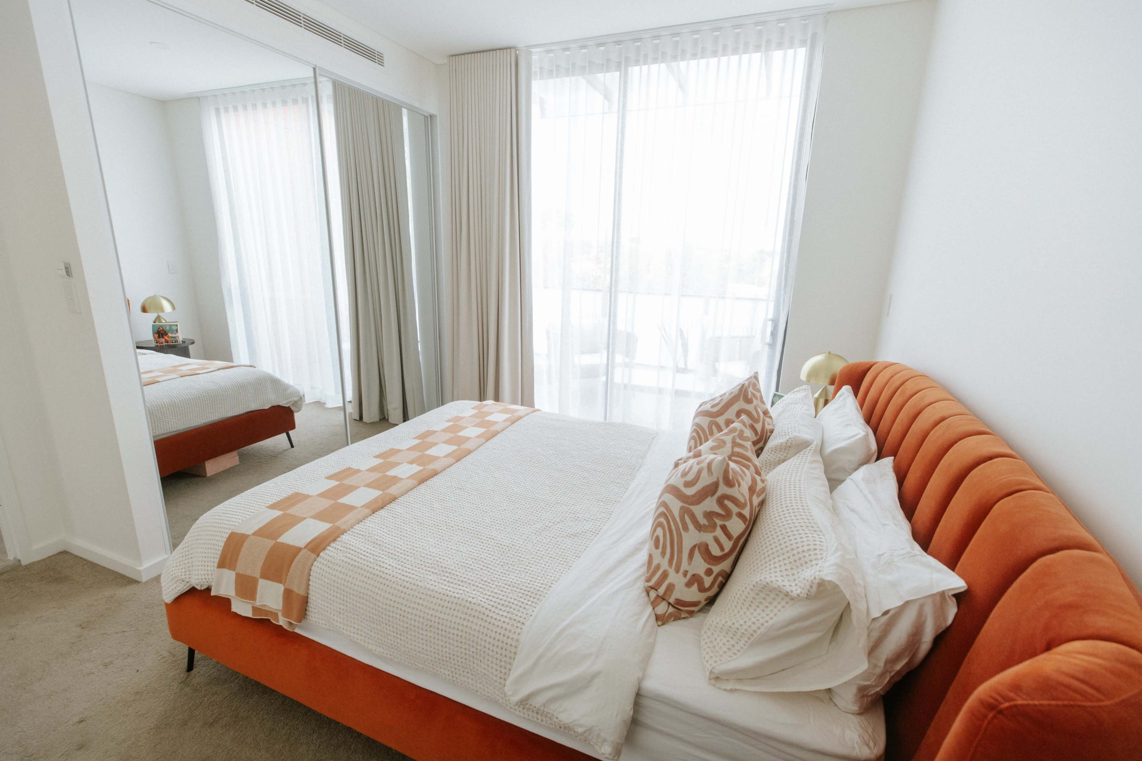 A well-lit bedroom featuring an orange upholstered bed with white bedding, decorative pillows, and a mirror reflecting part of the room.
