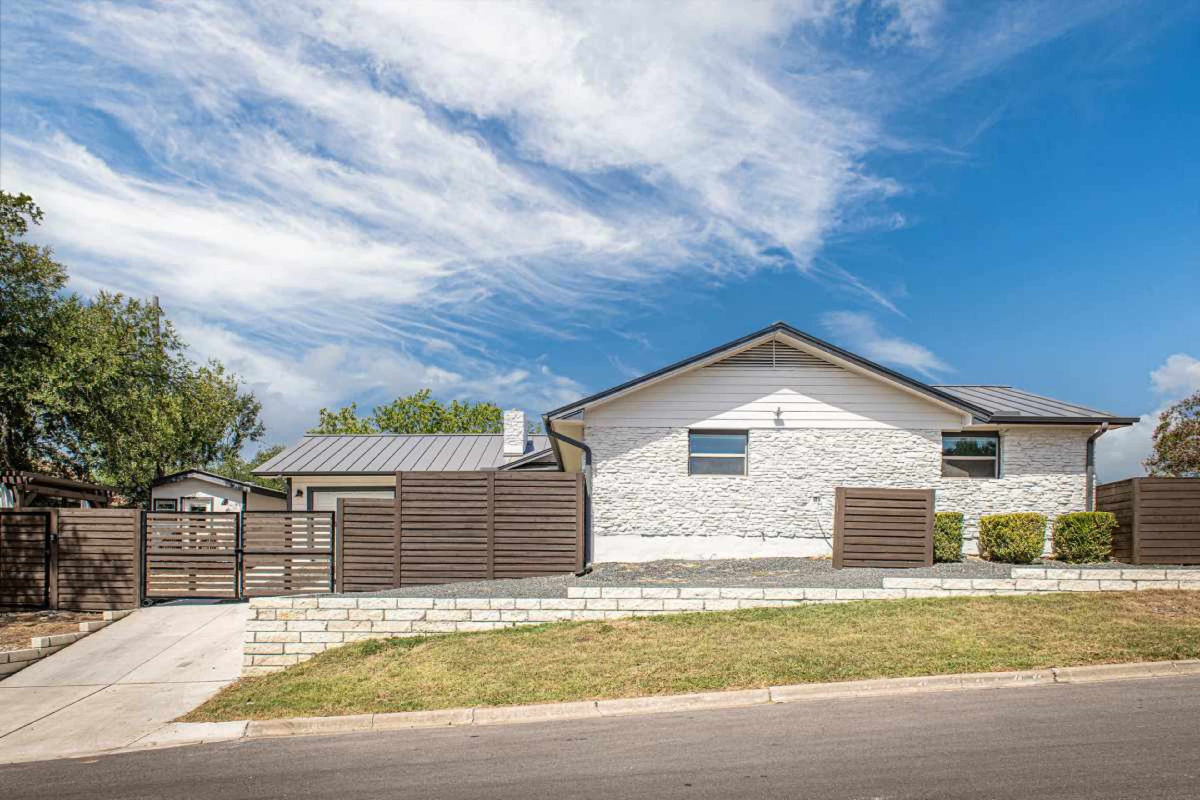 A modern white house with a stone exterior and a metal roof is set behind a wooden fence, surrounded by greenery and under a cloudy blue sky.