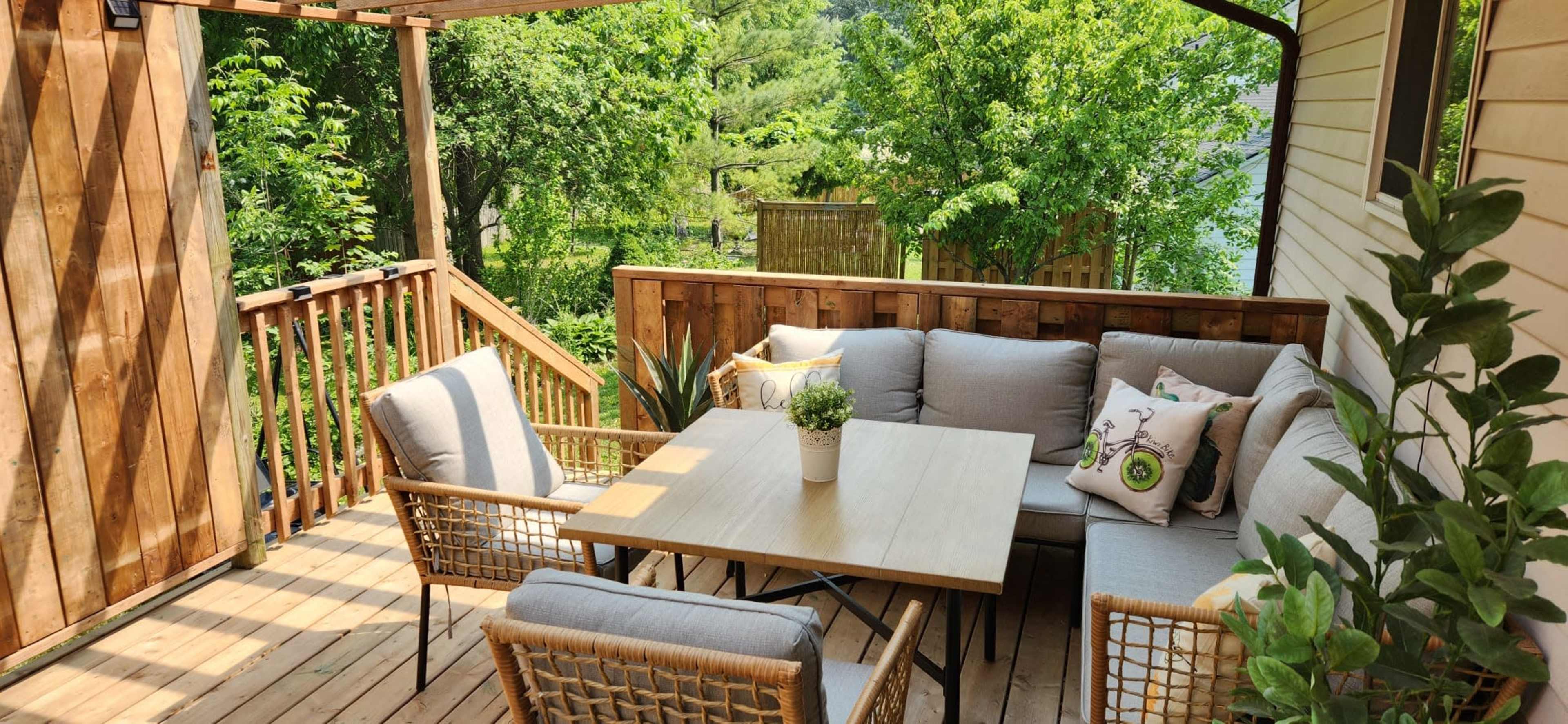 A wooden deck features a seating area with gray cushions, a light-colored table, and surrounding greenery.