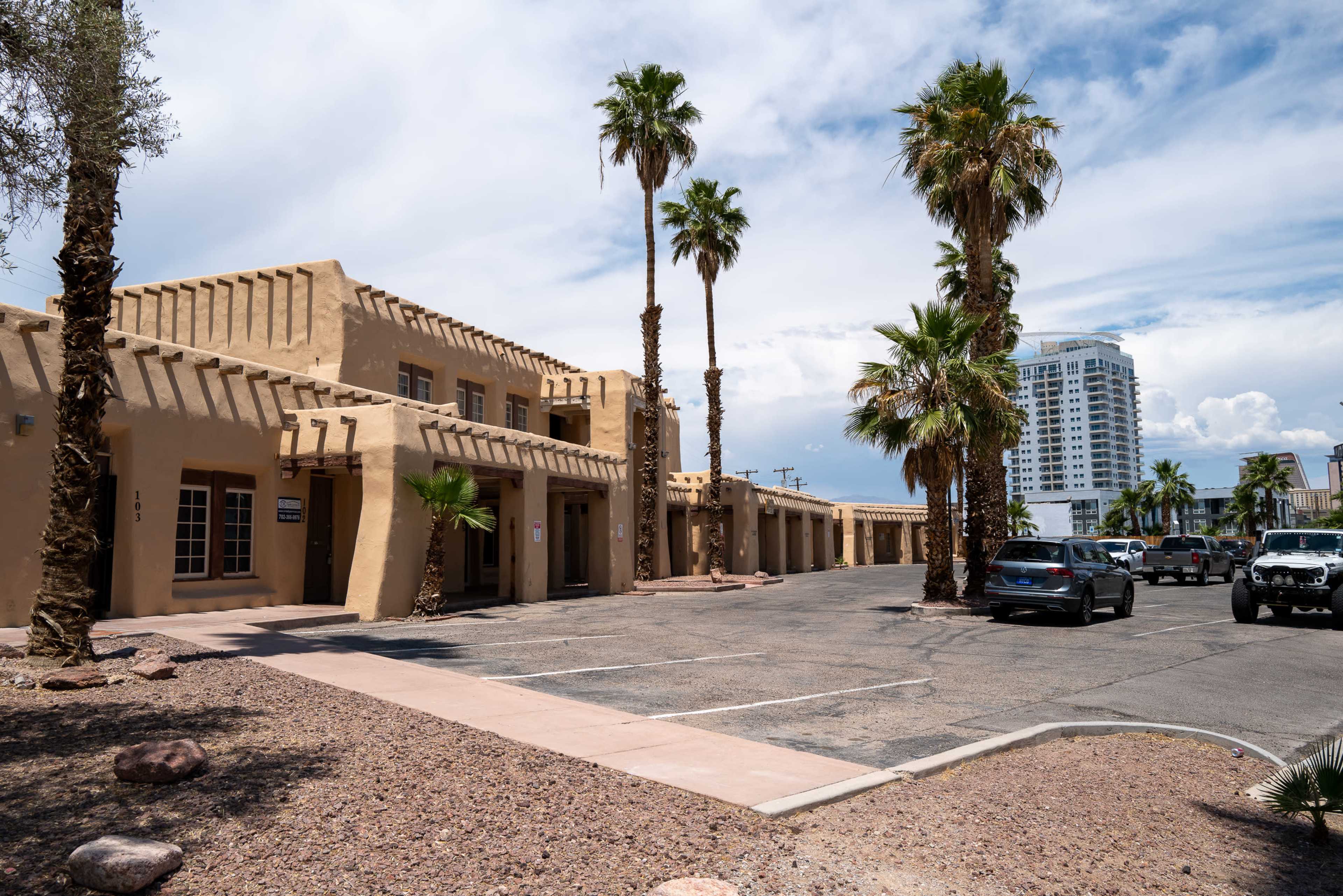 A low-rise adobe-style building surrounded by palm trees and a parking area, with a tall modern building visible in the background.