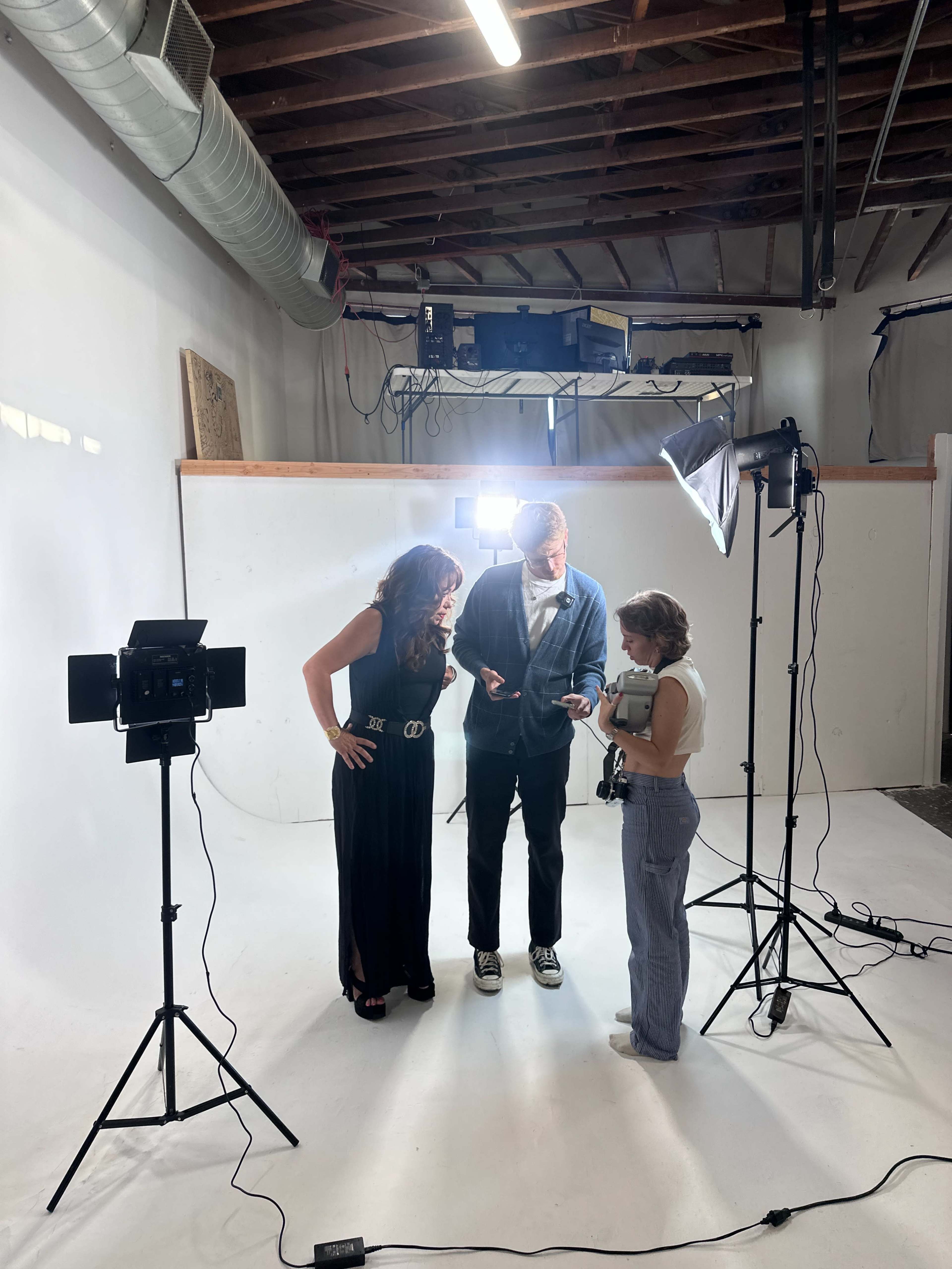 Three people are positioned in a photography studio with soft lighting, discussing something on a device while surrounded by camera equipment and a seamless backdrop.
