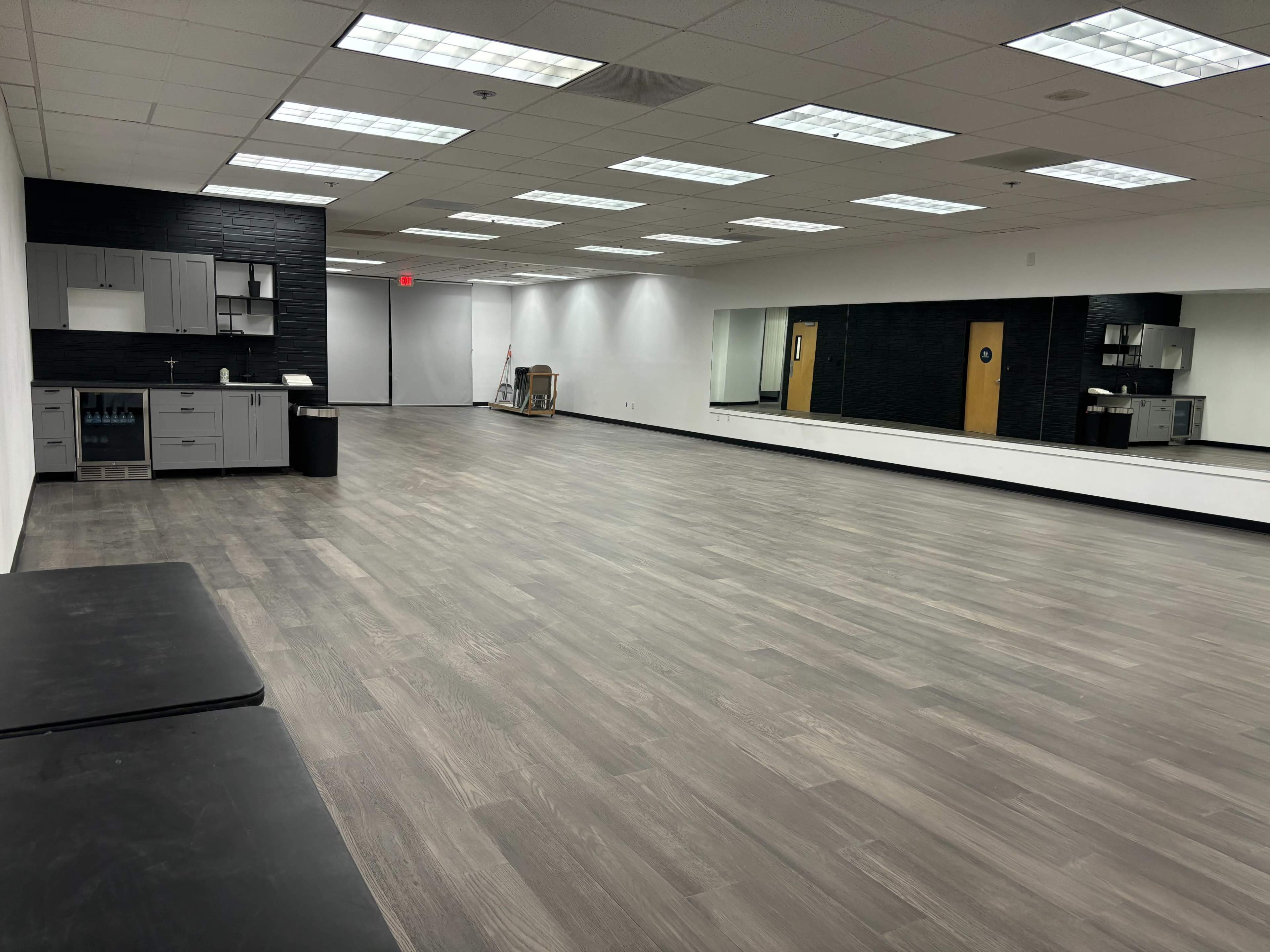 A spacious room with wooden flooring, equipped with a kitchenette on one side and a large mirror along the opposite wall.