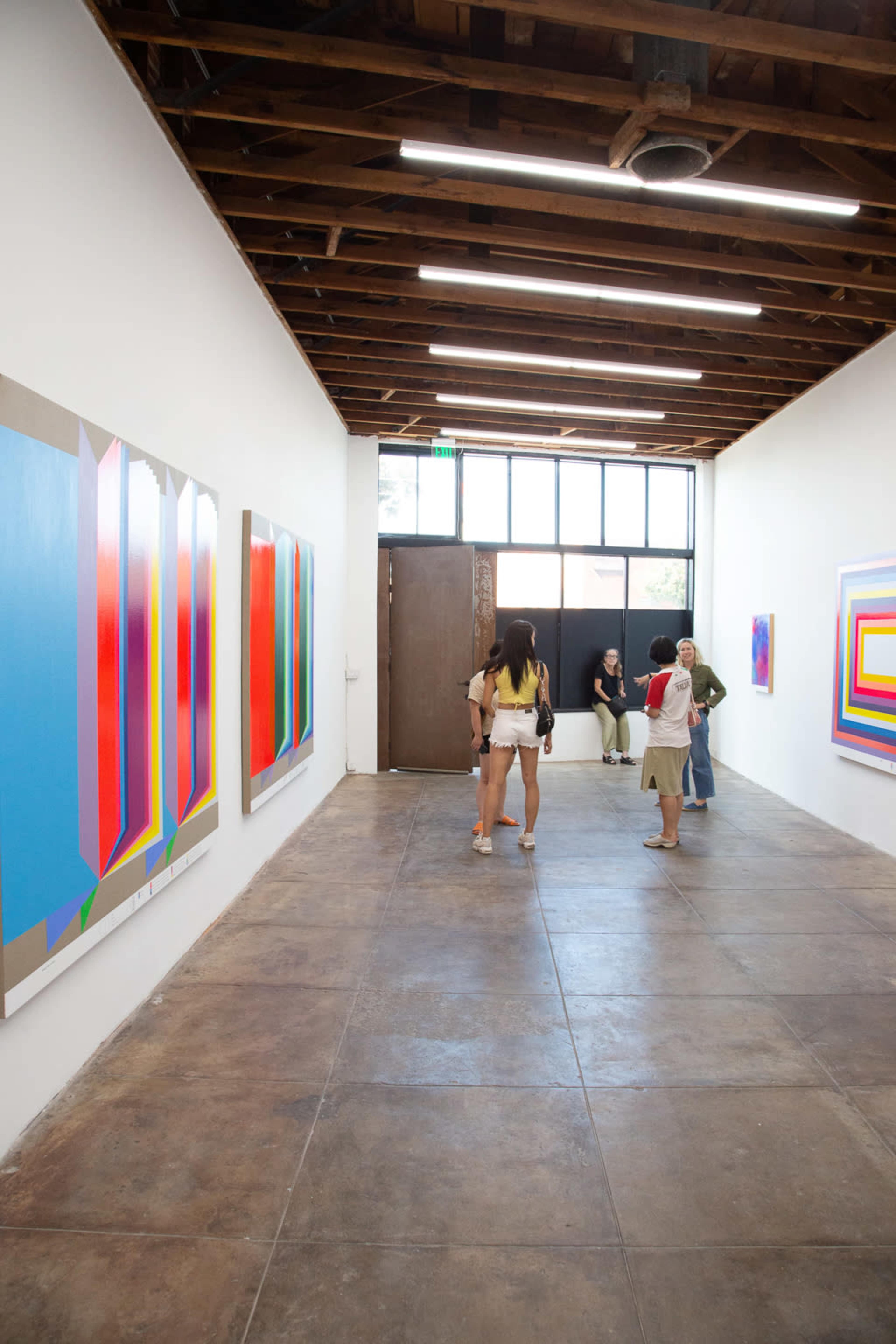 The image shows an art gallery with brightly colored abstract paintings on the walls and visitors engaged in conversation near the entrance.