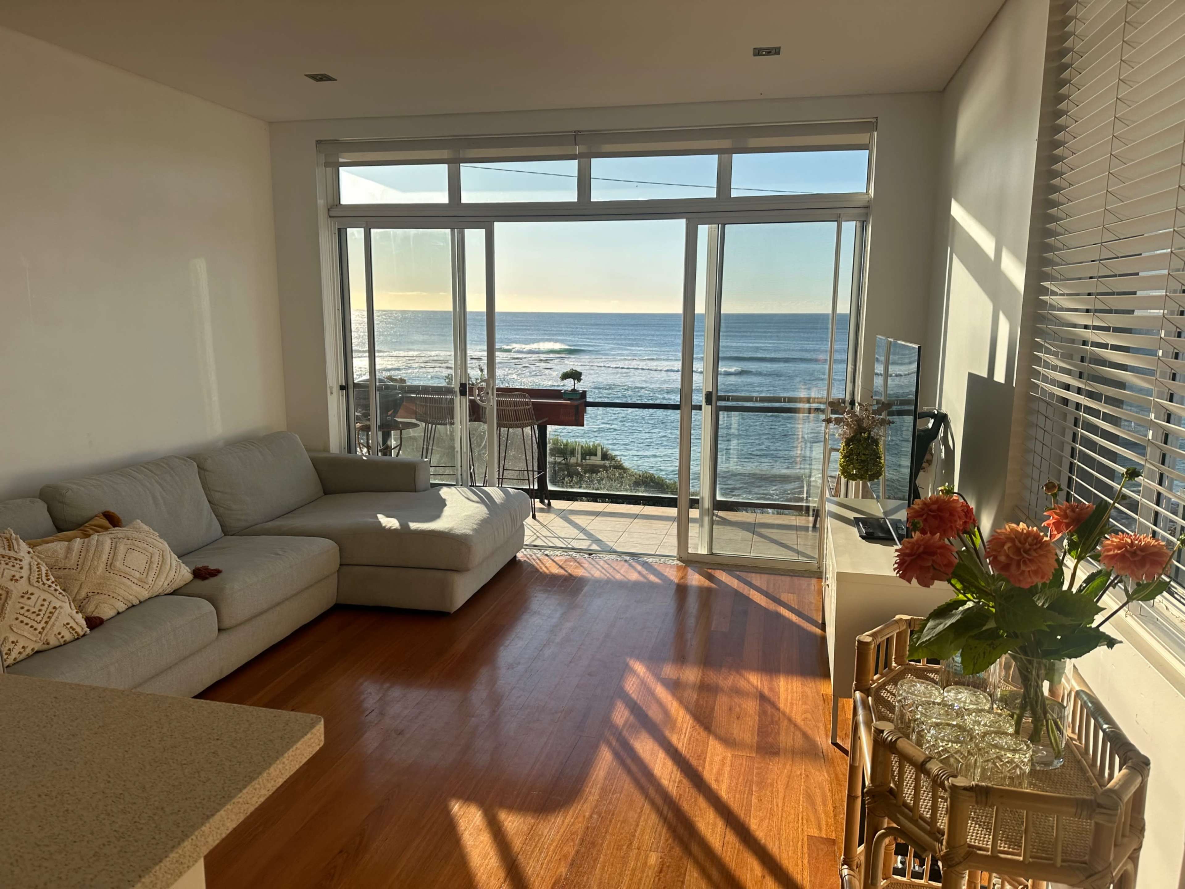 A living room features a gray sofa, a coffee table with flowers, and large windows overlooking the ocean.
