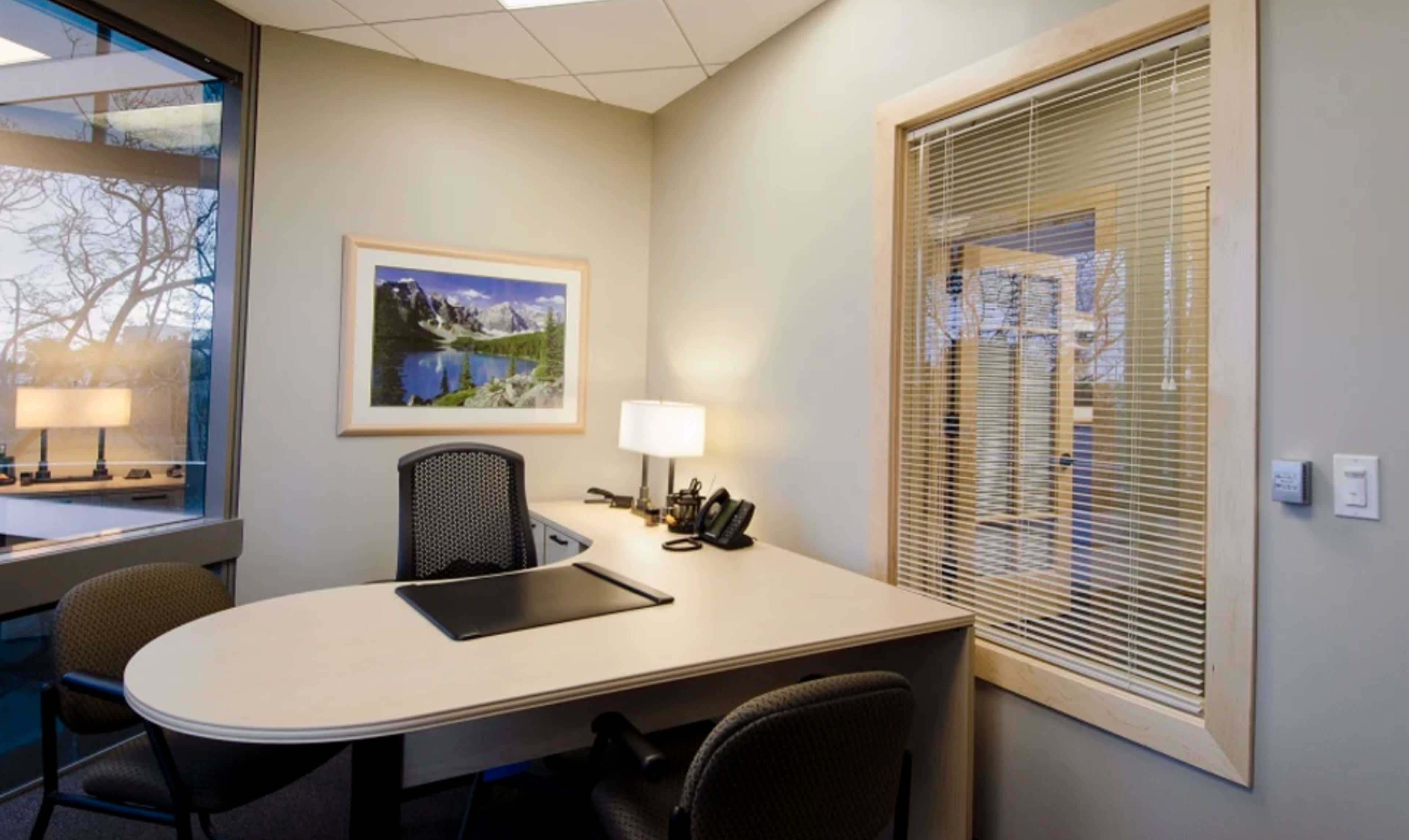 The image shows a small office with a desk, two chairs, a lamp, and a large window featuring a scenic landscape photograph on the wall.