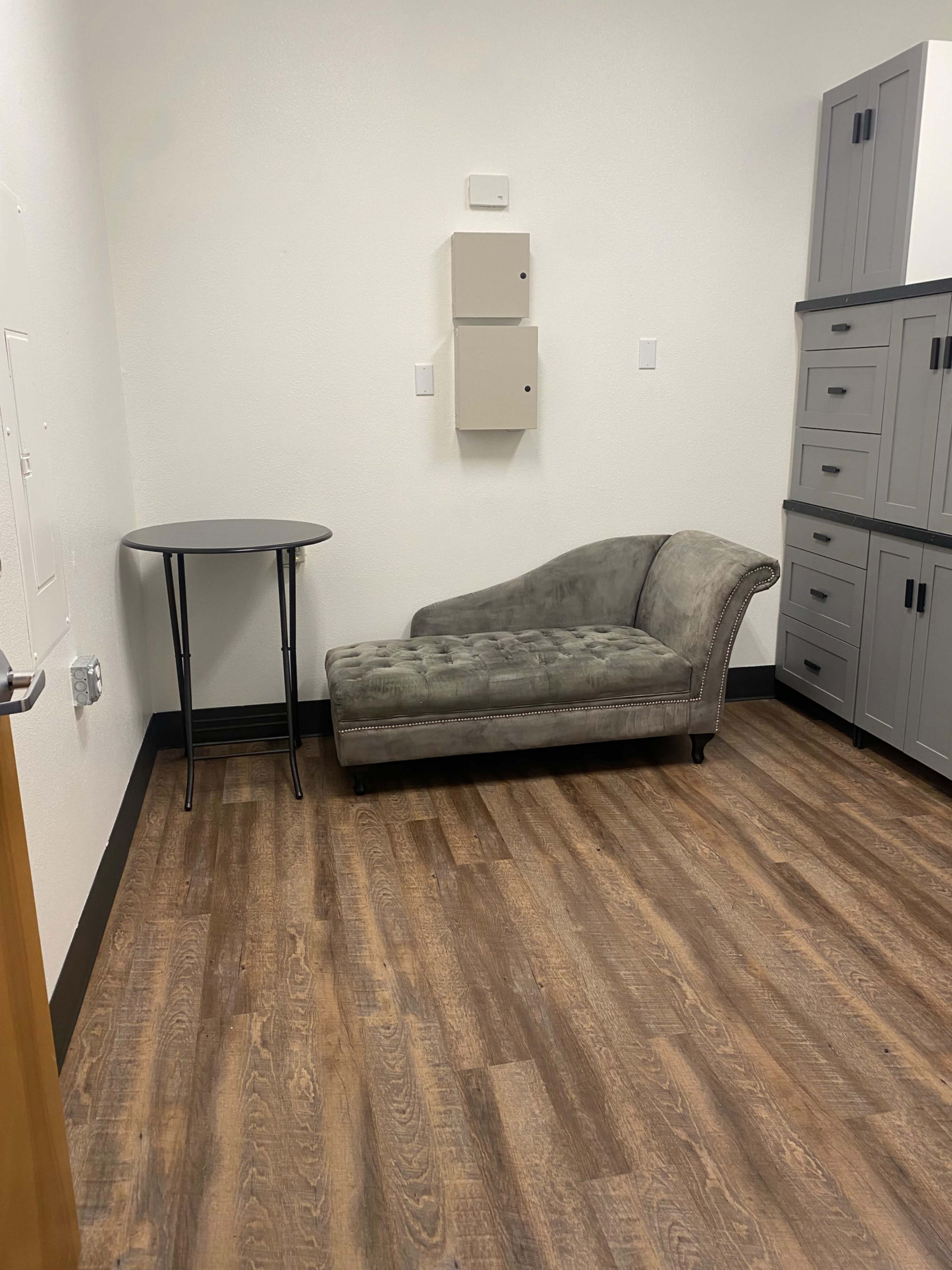 The image shows a small room with a gray chaise lounge, a round table, and gray cabinets against the wall.