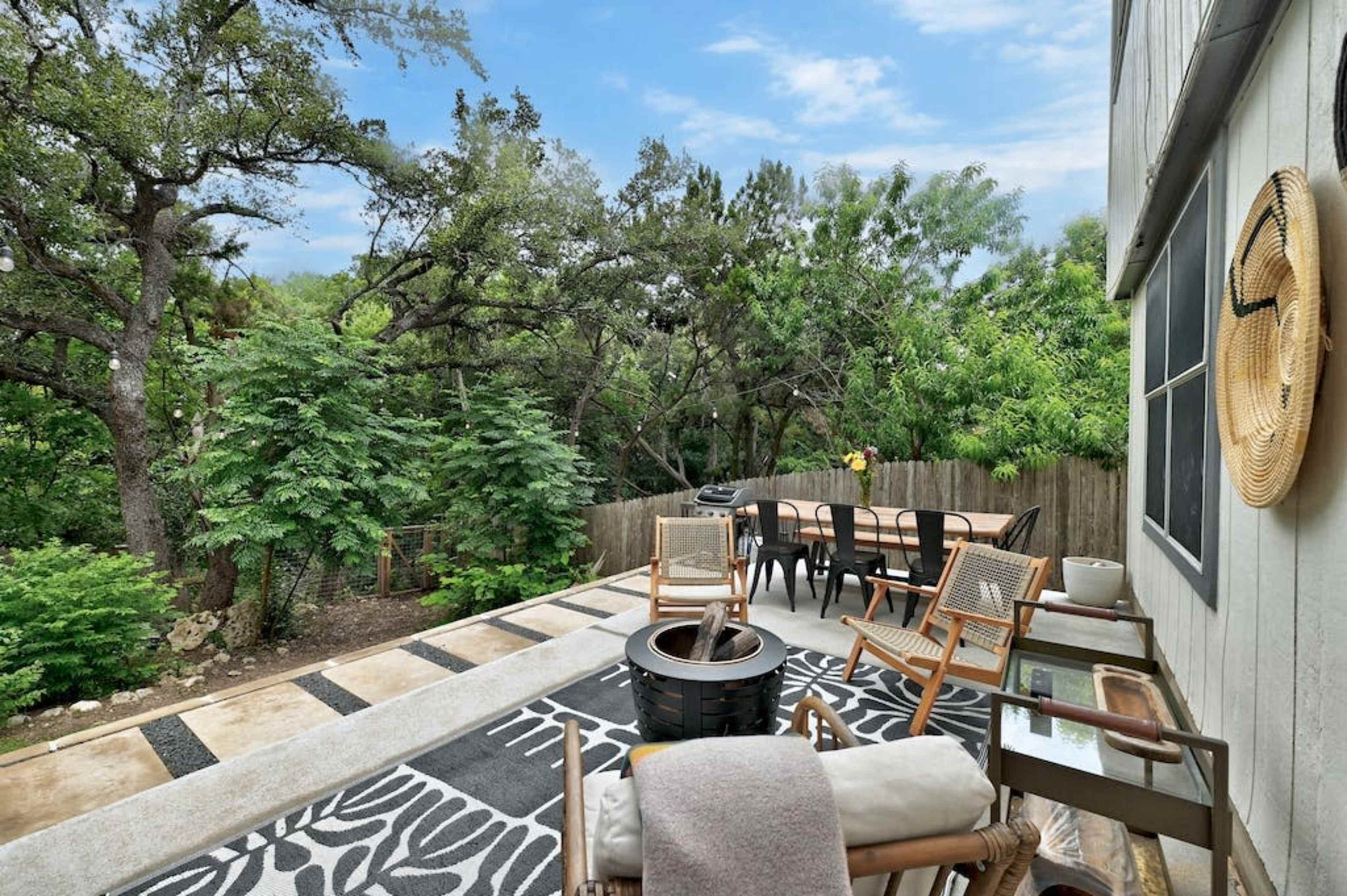 The image shows a patio area with seating, a dining table, and a view of trees and greenery in the background.