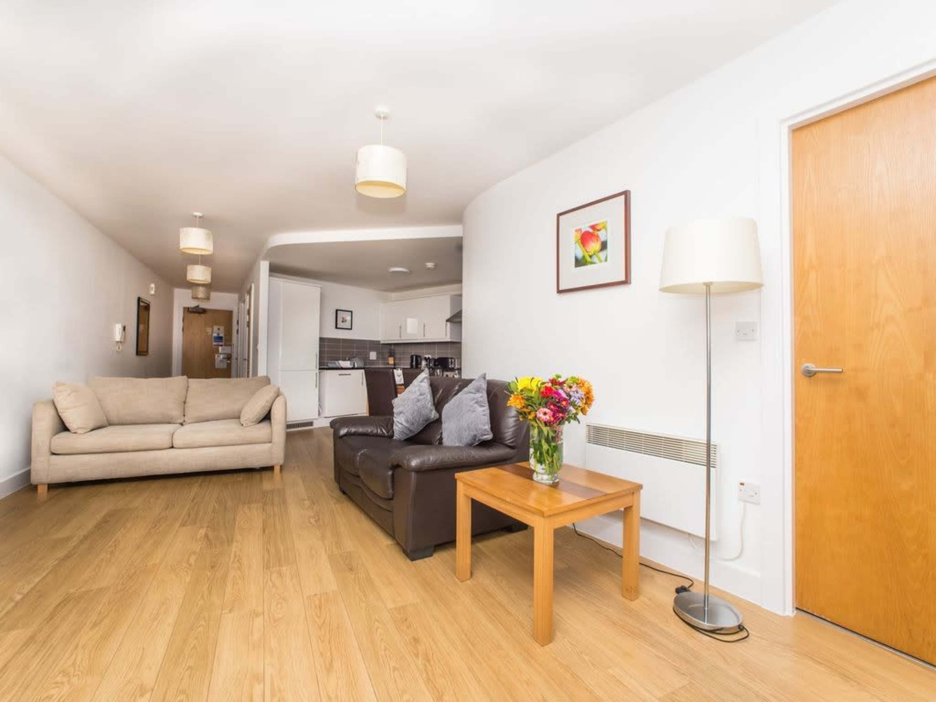 A living room with a brown couch, a wooden coffee table with a vase of flowers, and a light-colored wall leading to a kitchen area in the background.