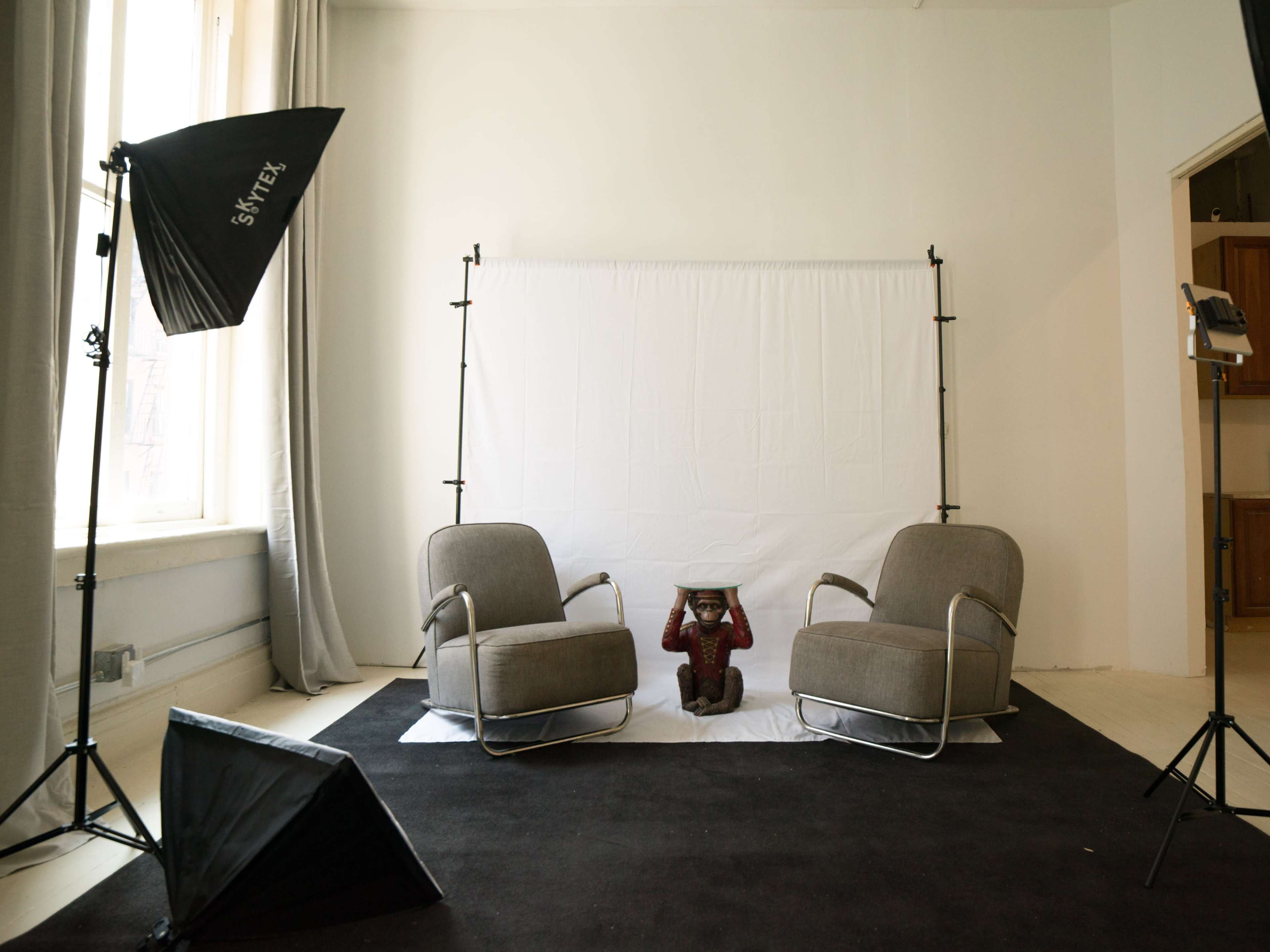 A minimal photography studio setup features two grey chairs, a black backdrop, a white screen, and lighting equipment.