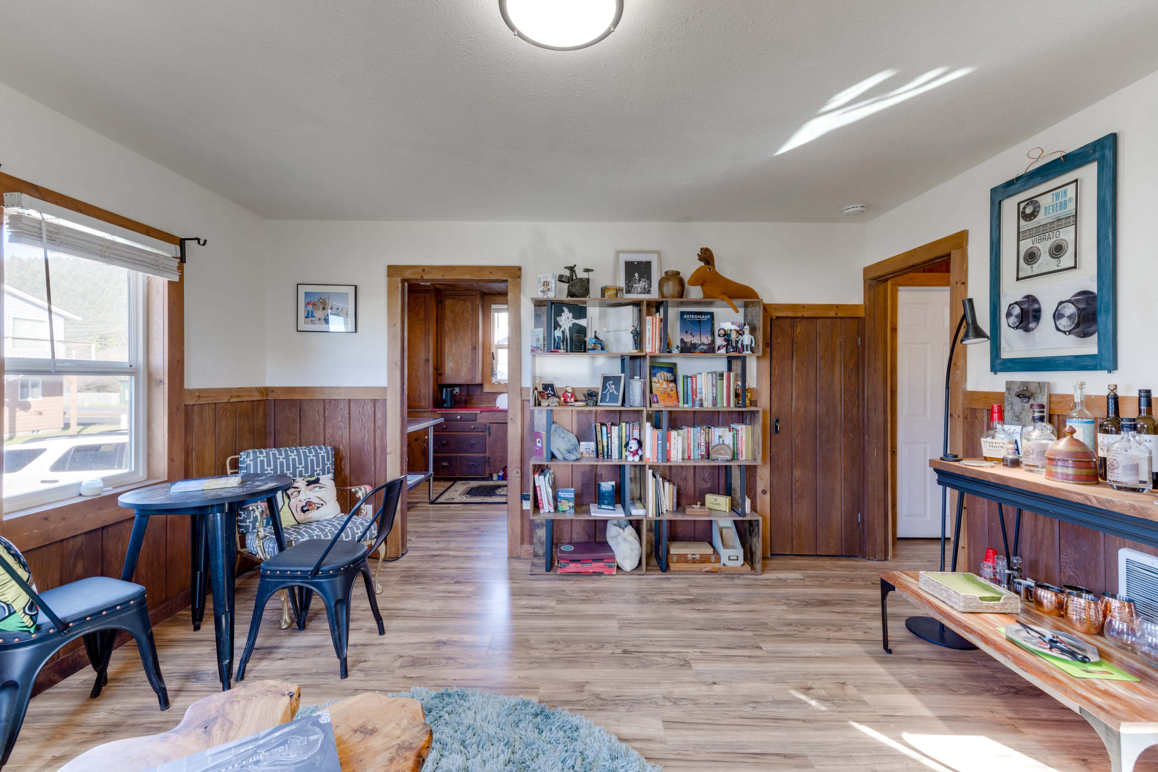 The image shows a cozy living room area with a wooden bookshelf, a small dining table set, and a kitchen visible in the background.