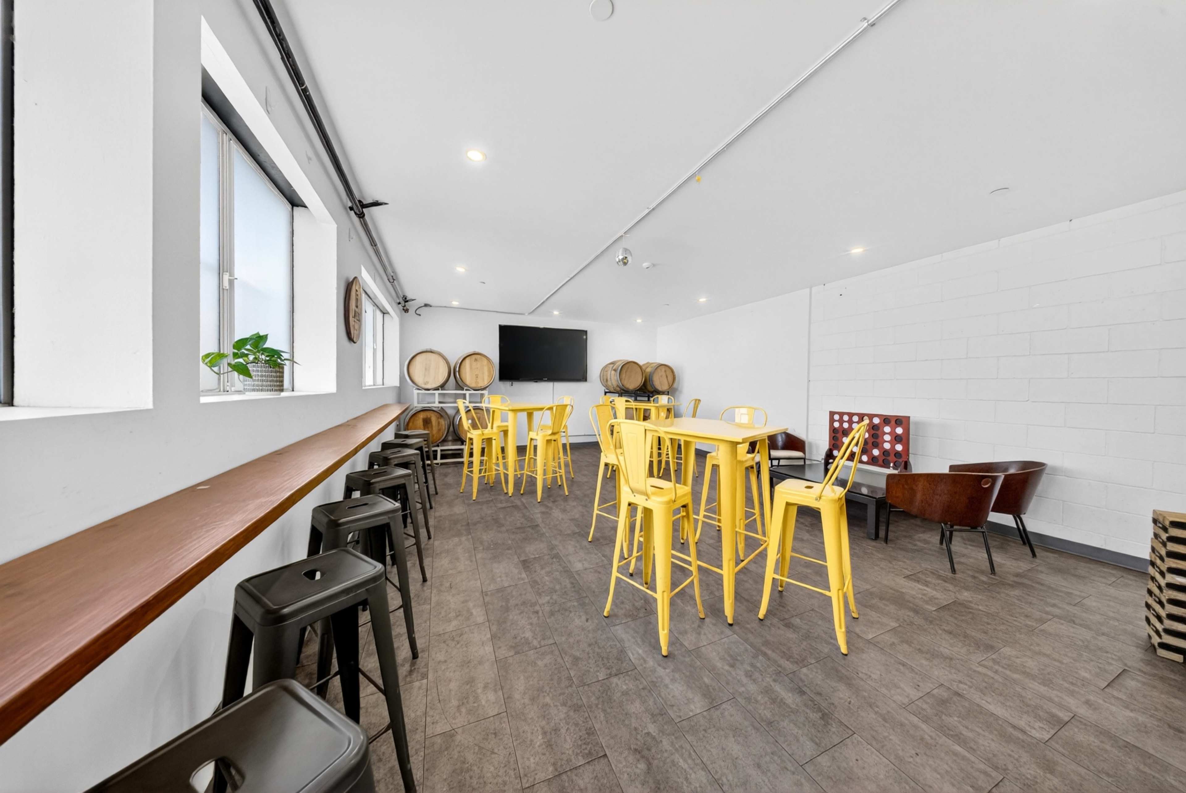 The image shows a spacious room with a mix of yellow and black bar stools and tall tables, wooden barrels in the background, and a large wall-mounted screen.
