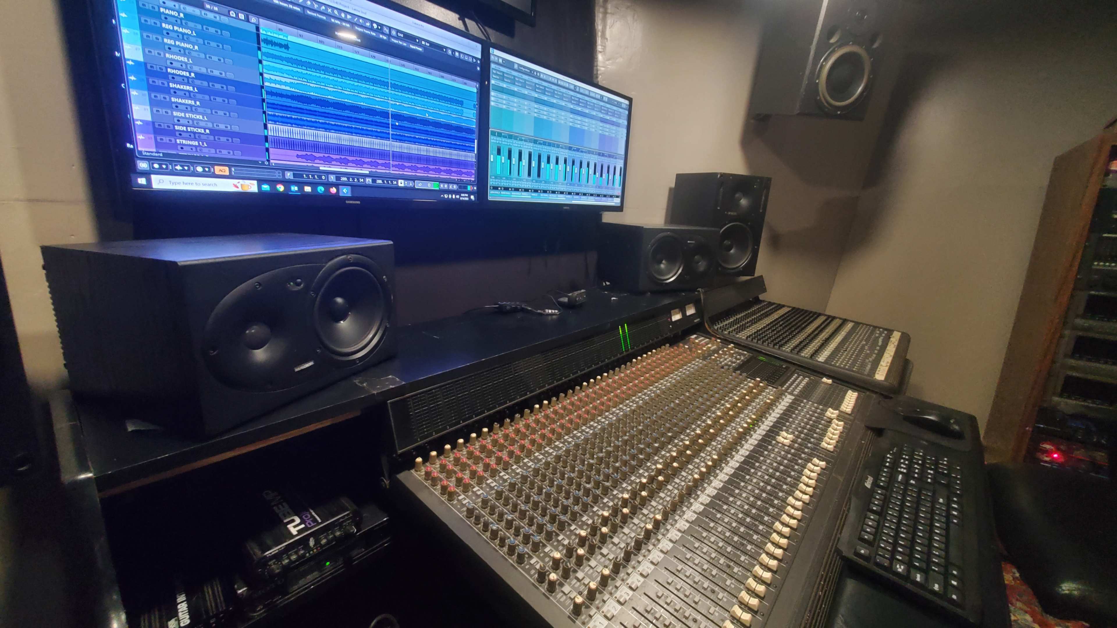 A music production studio featuring a large mixing console, two computer monitors displaying audio tracks, and studio speakers positioned on either side.