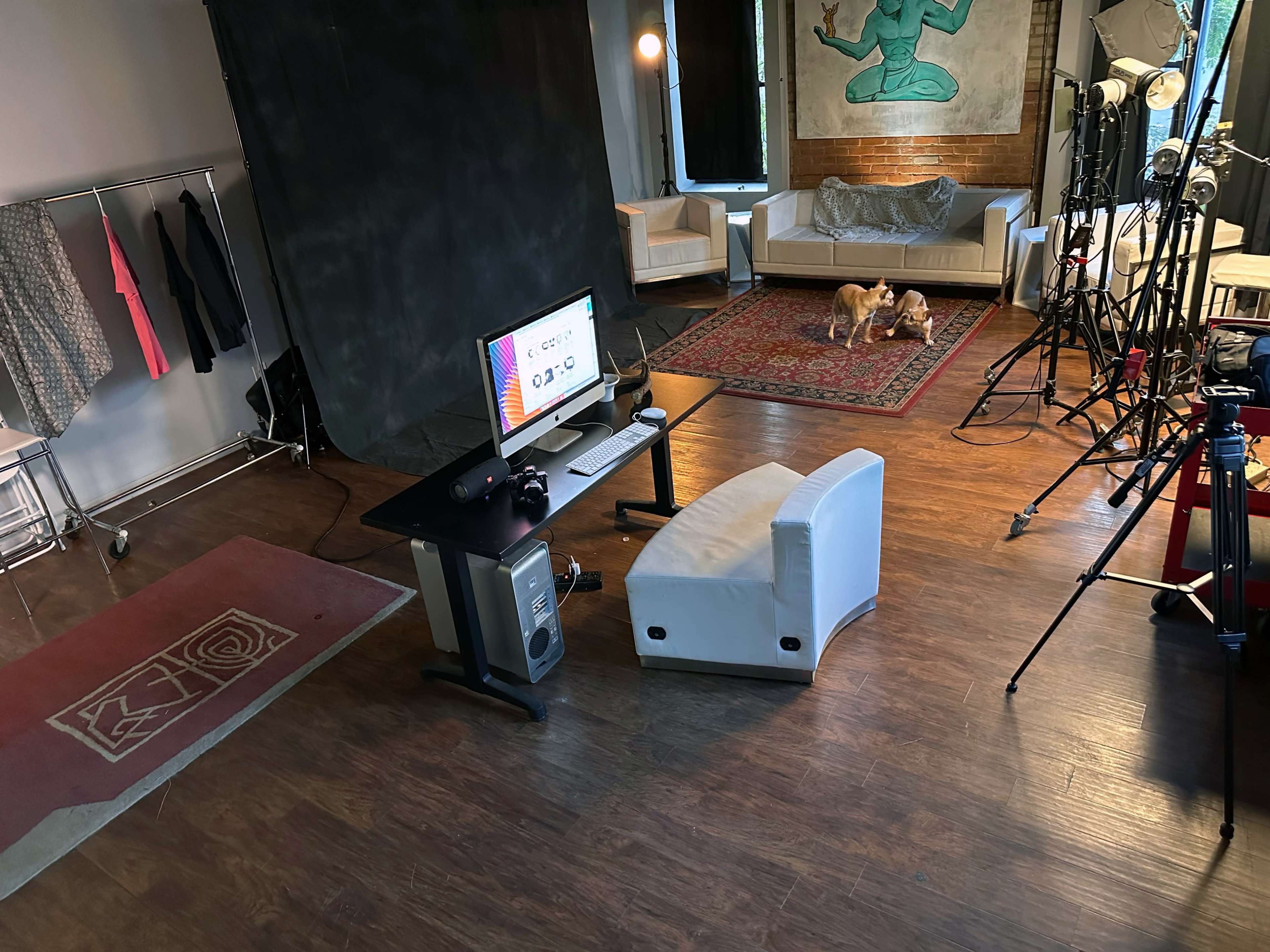 The image shows a studio space with a desk featuring a computer, a chair, and a backdrop alongside clothing racks, lighting equipment, and a couch.