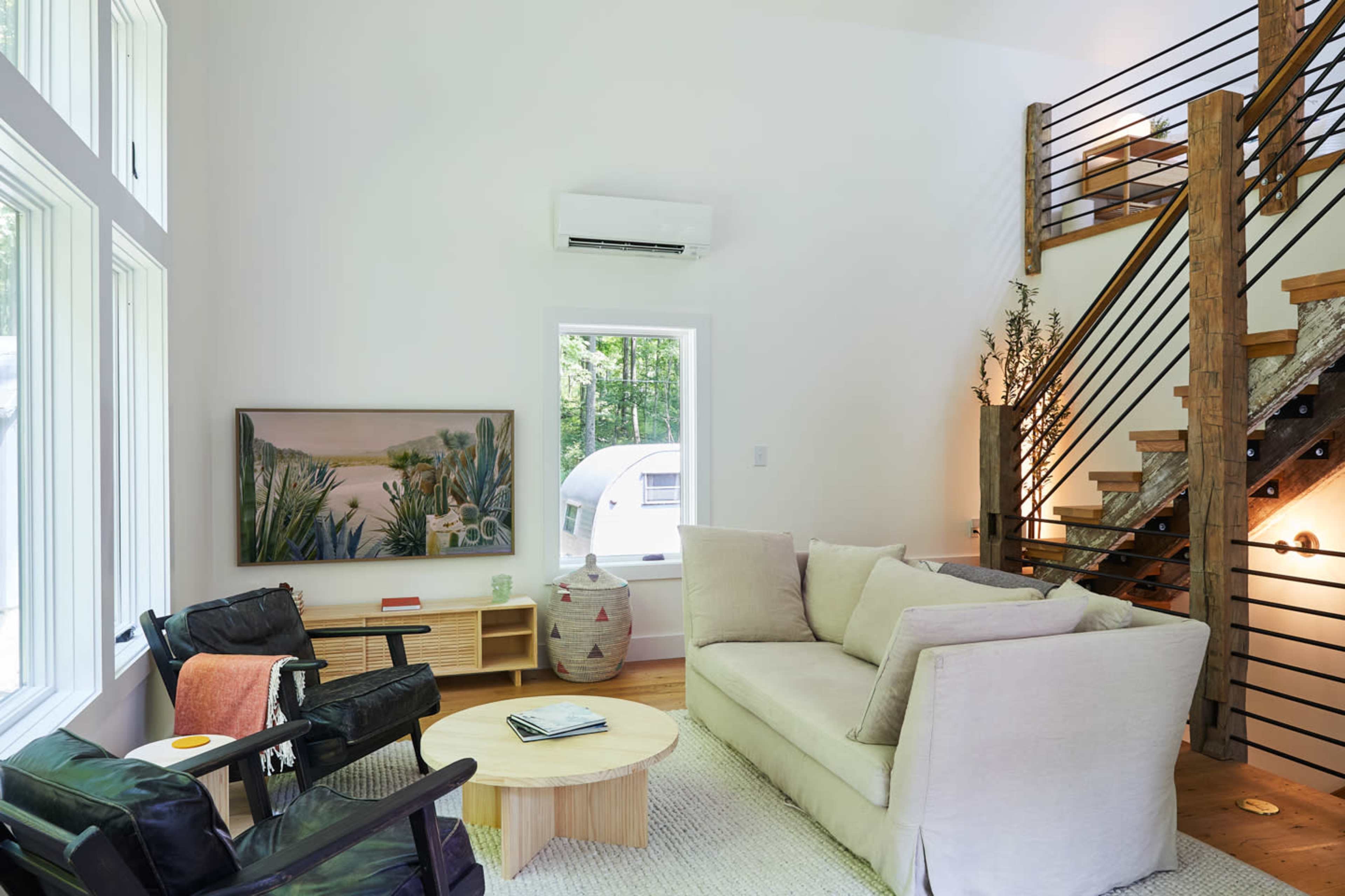 A modern living room with a beige sofa, black armchairs, a wooden coffee table, and a staircase with metal railings, all placed next to large windows allowing natural light.
