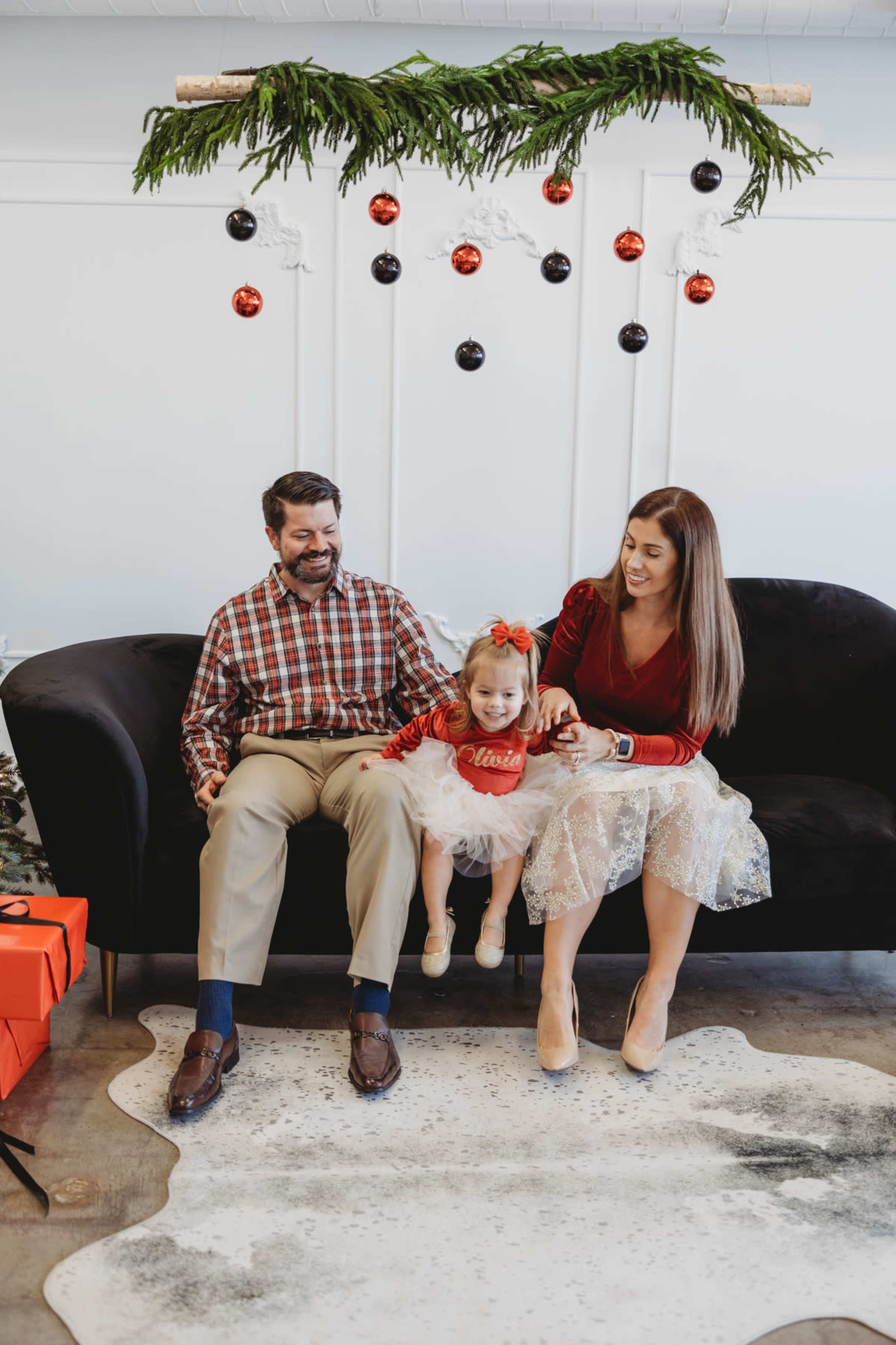 A family sits together on a black couch decorated for the holidays, with the mother and father playfully interacting with their young daughter, who is wearing a festive outfit.