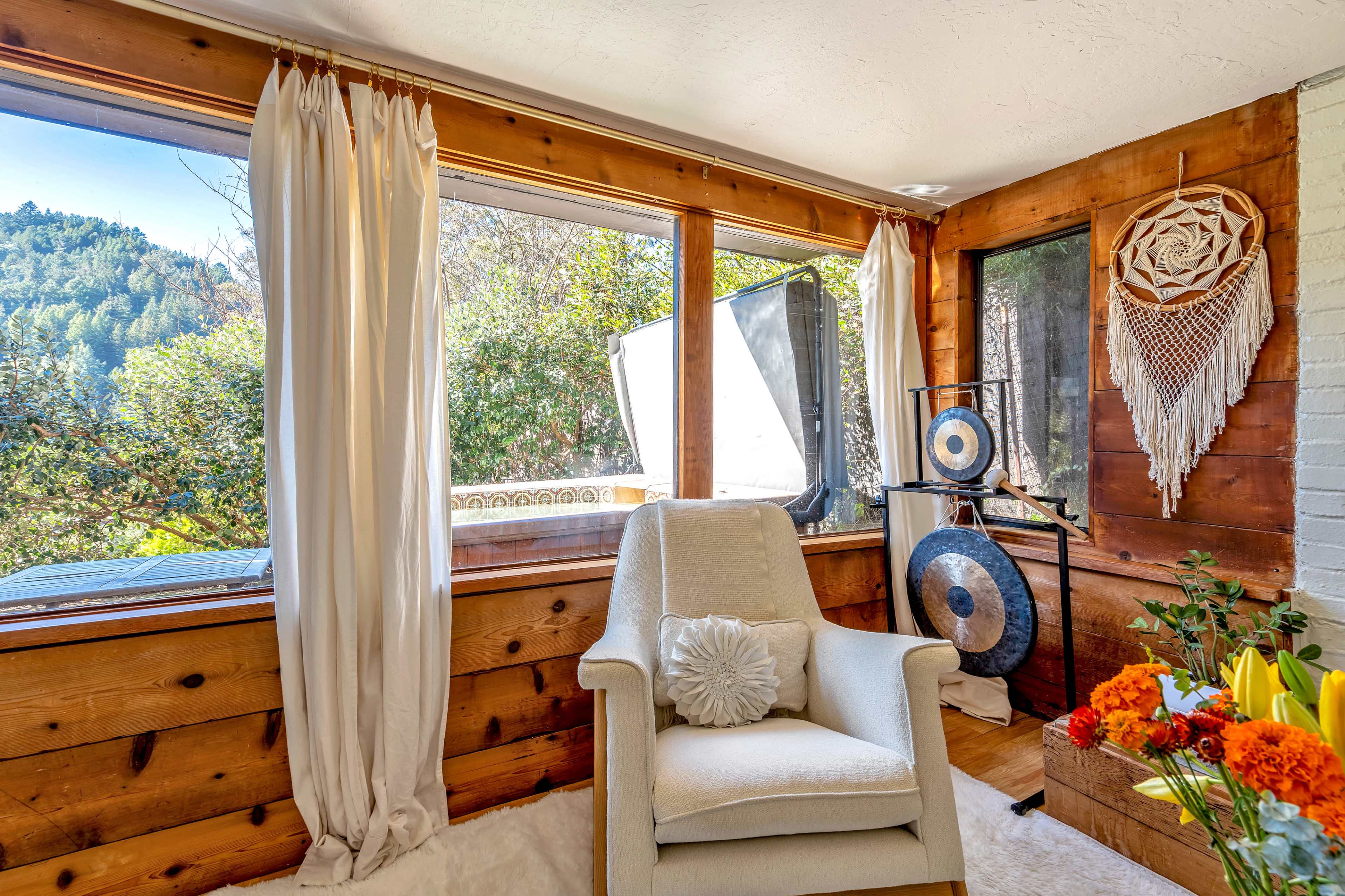 A bright room features a white armchair, large windows with views of trees, and decorative elements including a wall hanging and a plant.
