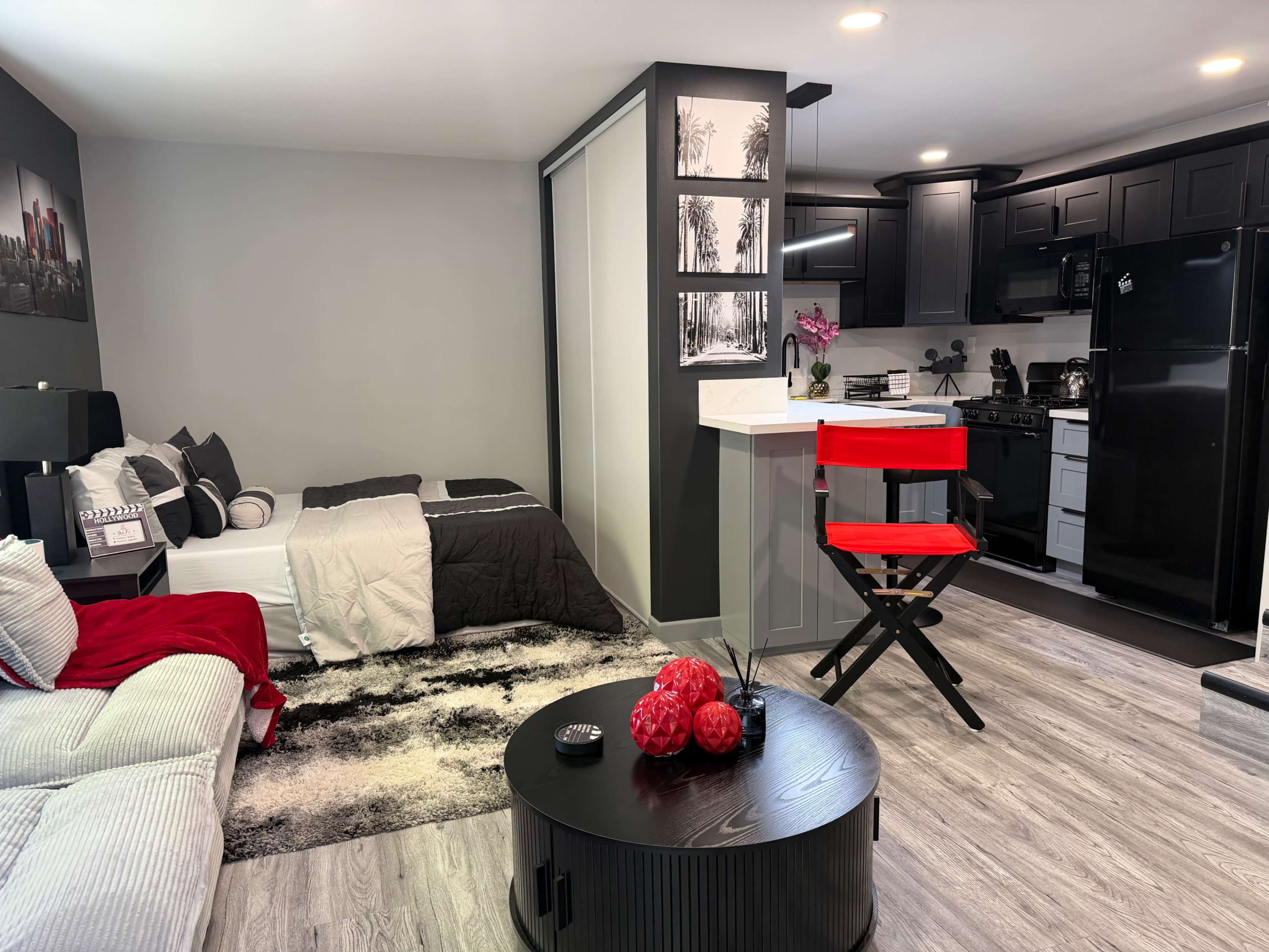 A small studio apartment features a bed with gray bedding, a kitchenette with black appliances, a red director's chair, and a round black coffee table decorated with red decor.