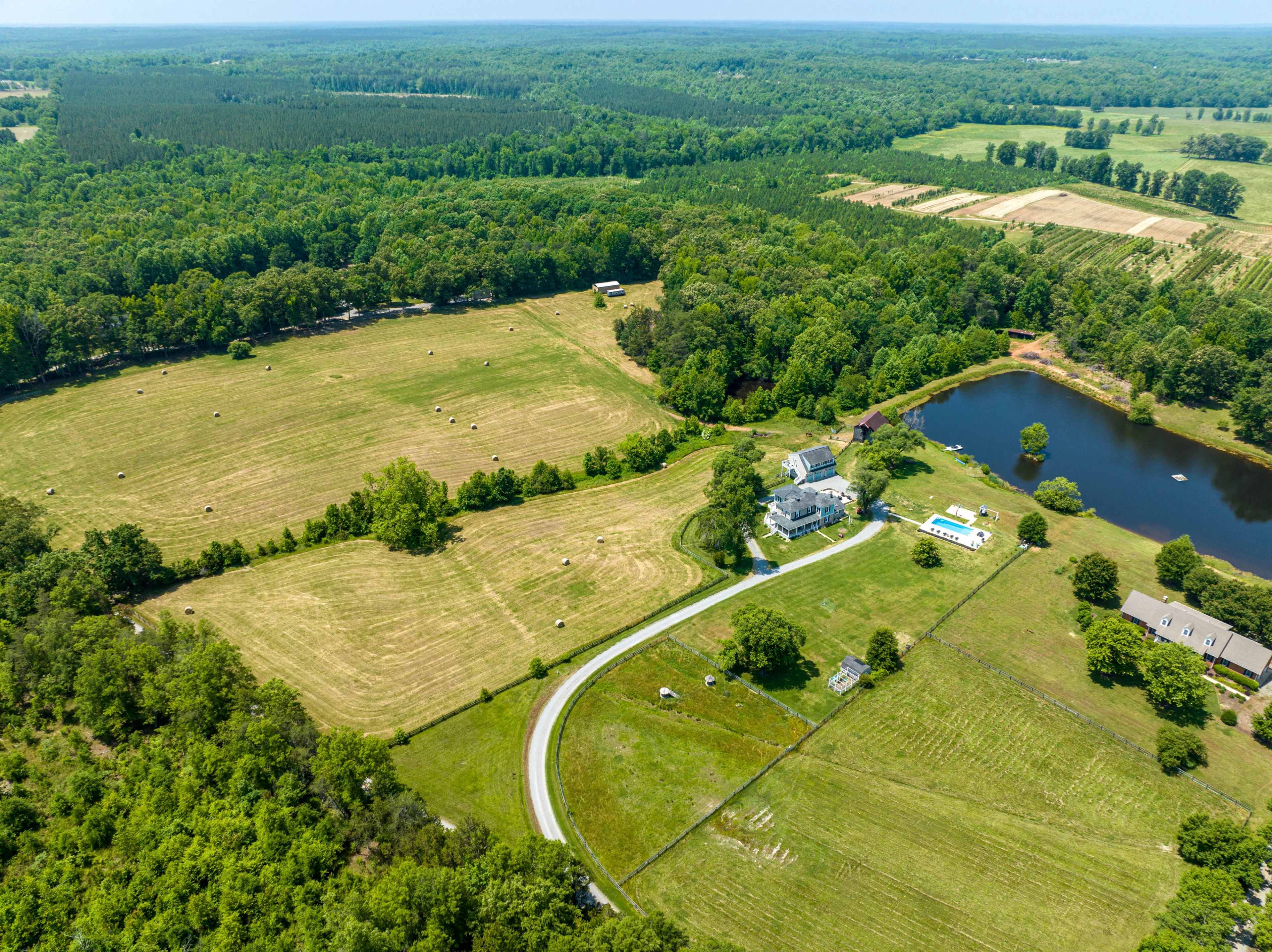 The aerial view shows a spacious rural landscape featuring a house near a pond, surrounded by fields and wooded areas.