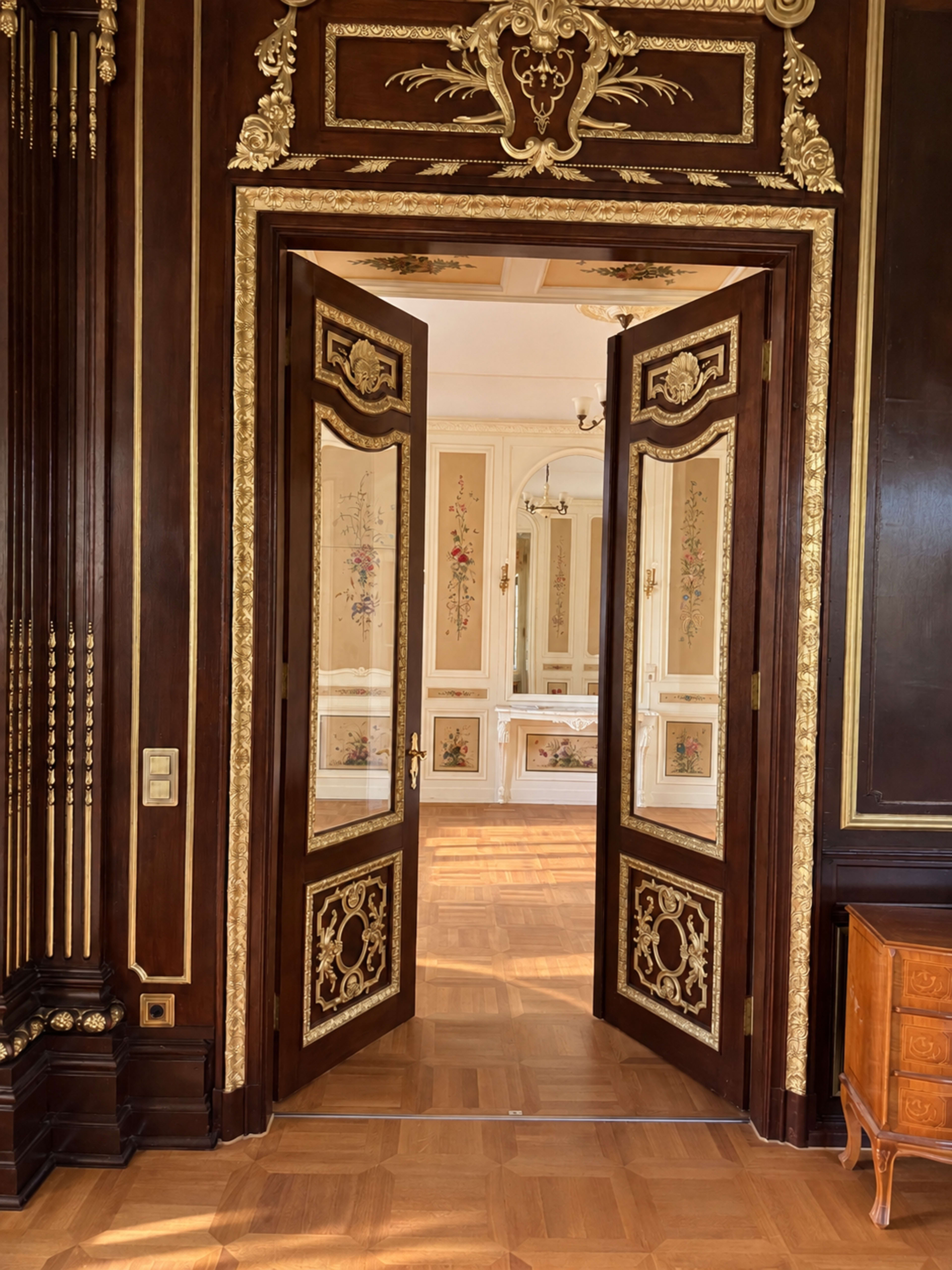 The image shows a pair of ornate wooden doors opening into a lavishly decorated room with intricate gold detailing and floral motifs on the walls.