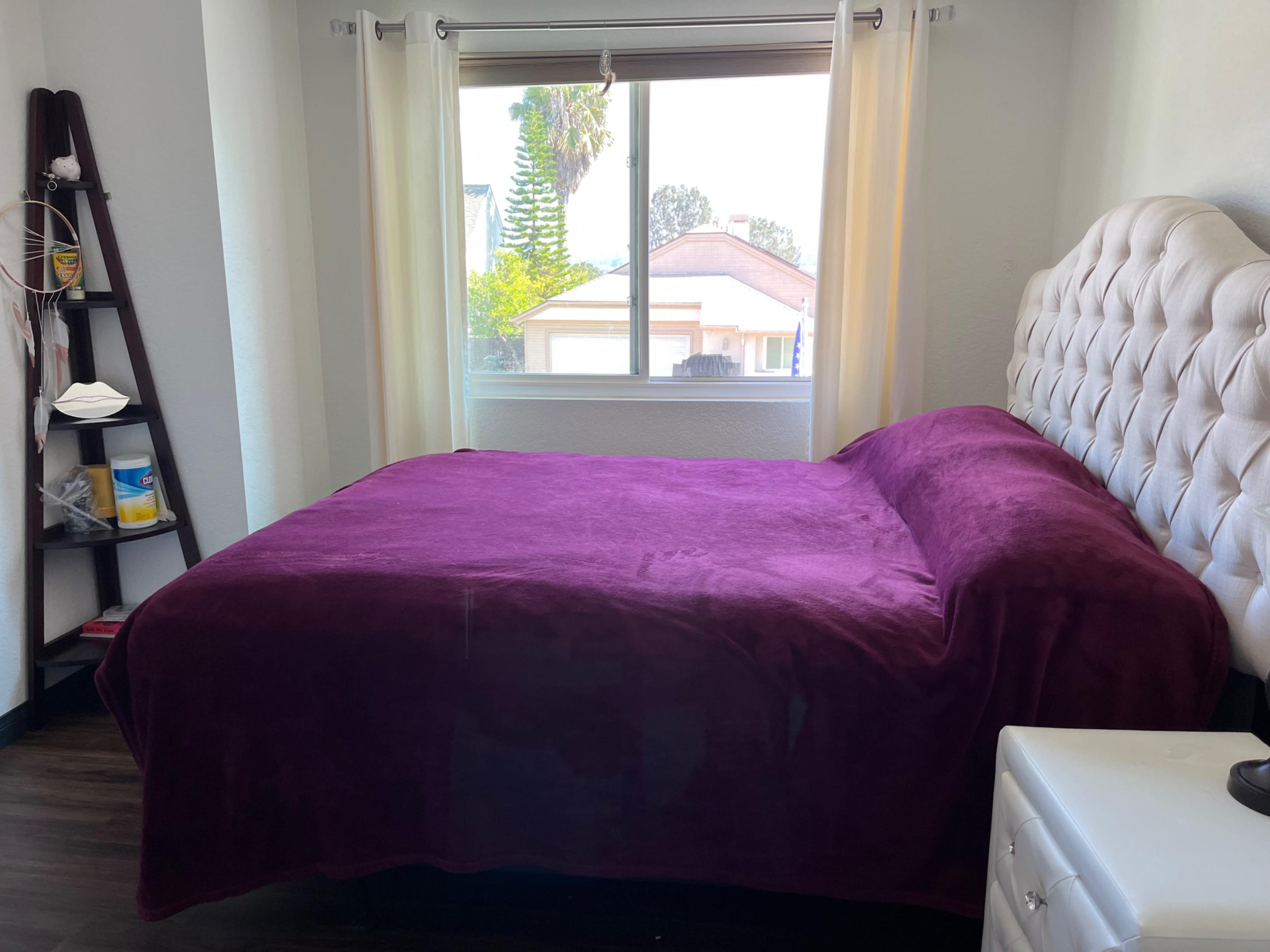 A bedroom features a bed with a purple blanket, a window with sheer curtains, and a small bedside table beside it.