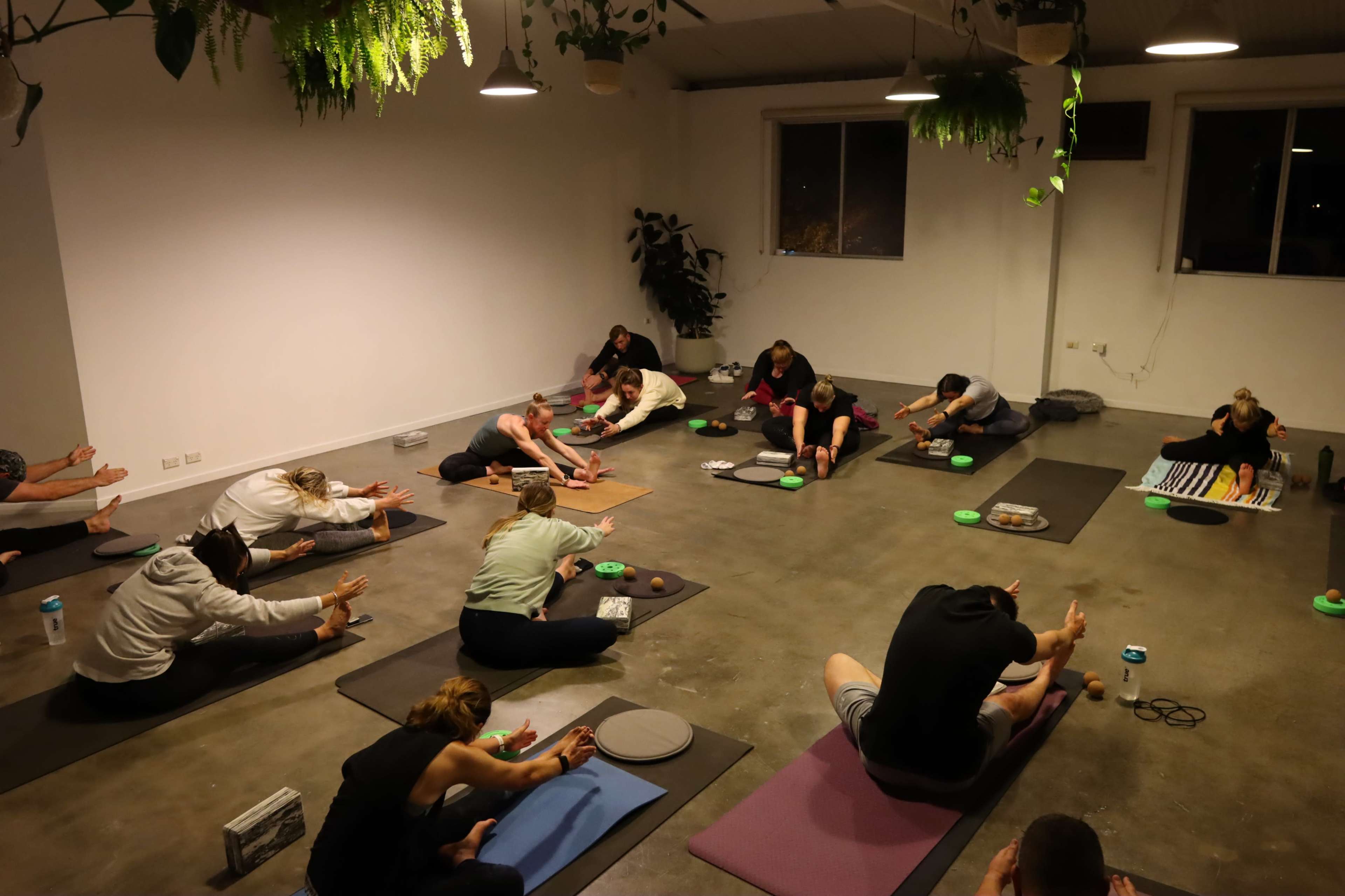 A group of people practice yoga in a spacious, dimly lit studio with mats and props scattered around.
